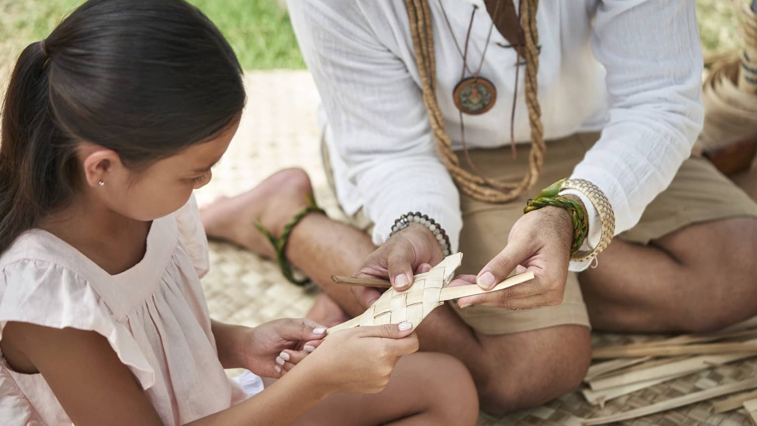 An adult shows a young girl how to weave a basket