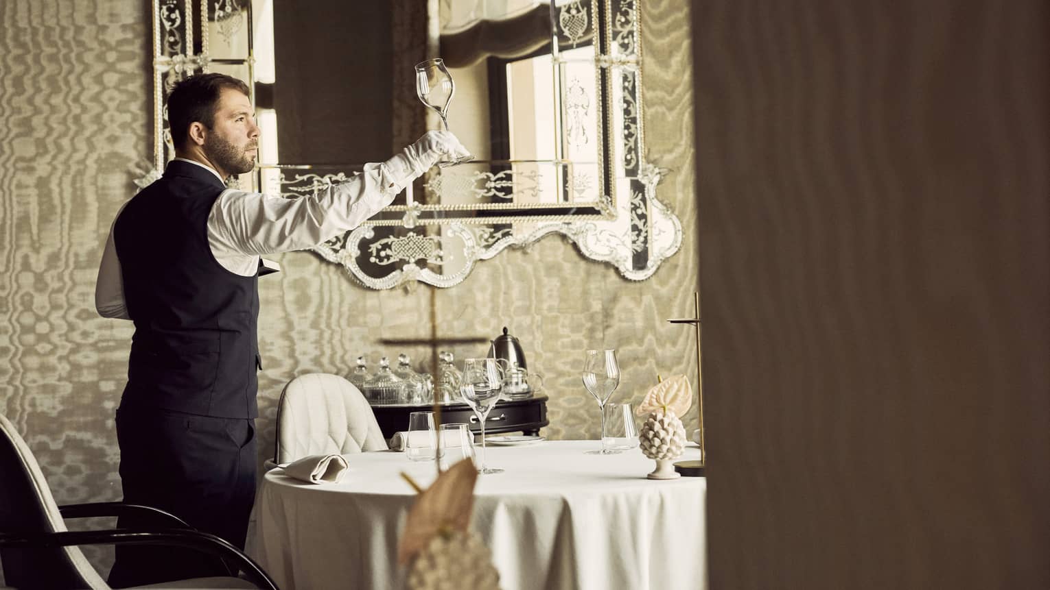 Server wearing black pants, black vest, white long-sleeve button down shirt and white gloves inspects an empty wine glass in a dining room with a large ornate mirror behind them