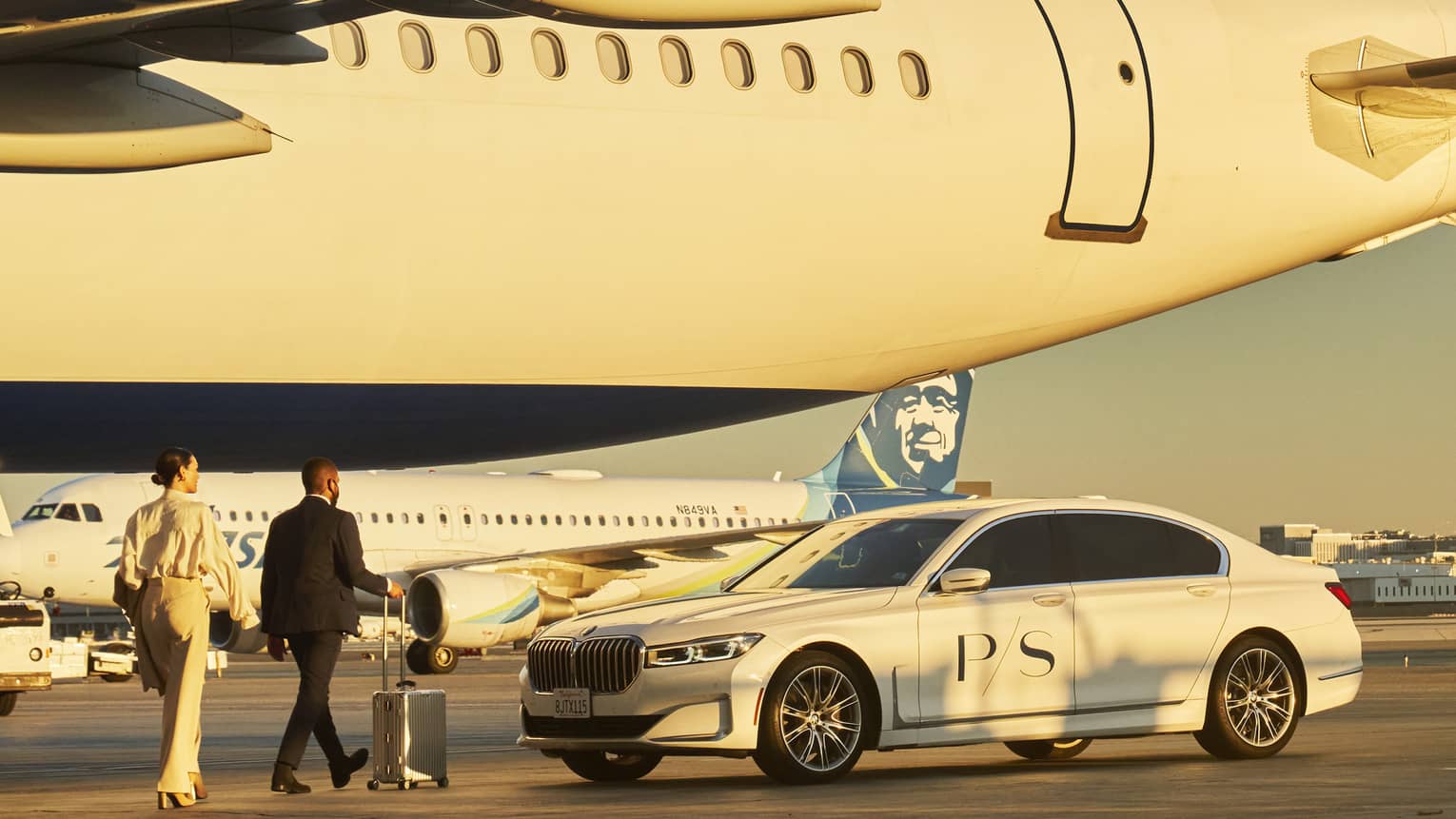 Two people walking toward airplane on tarmac, with a car in the foreground and another airplane in the background