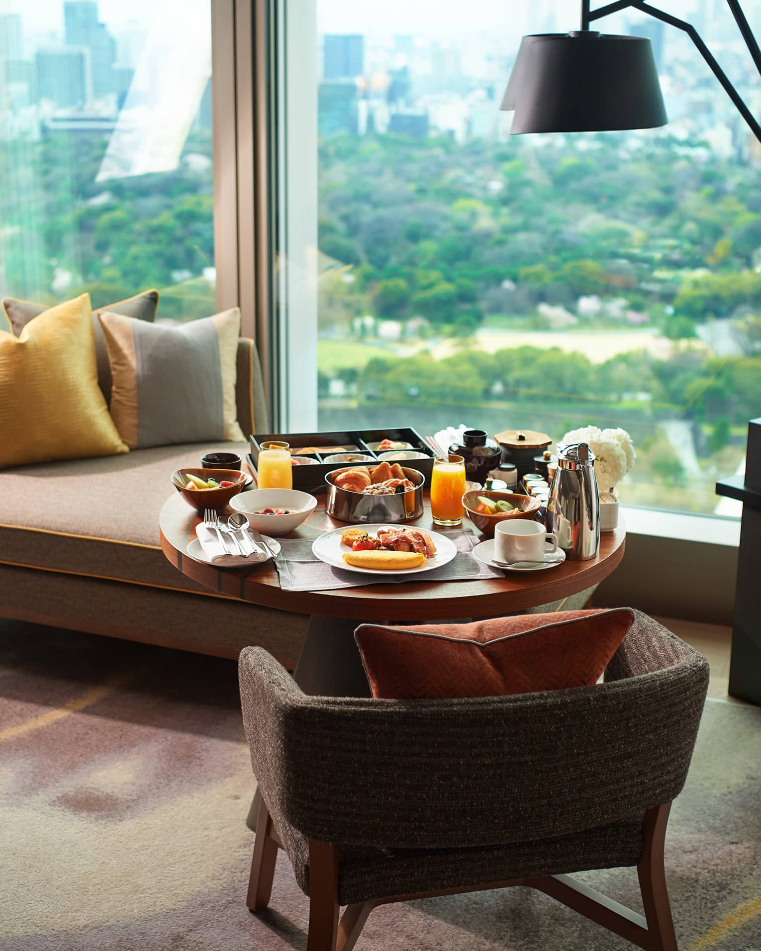 A breakfast tray of pastries, fresh fruit, juice and coffee on a guest room lounge chair at a window overlooking lush gardens.