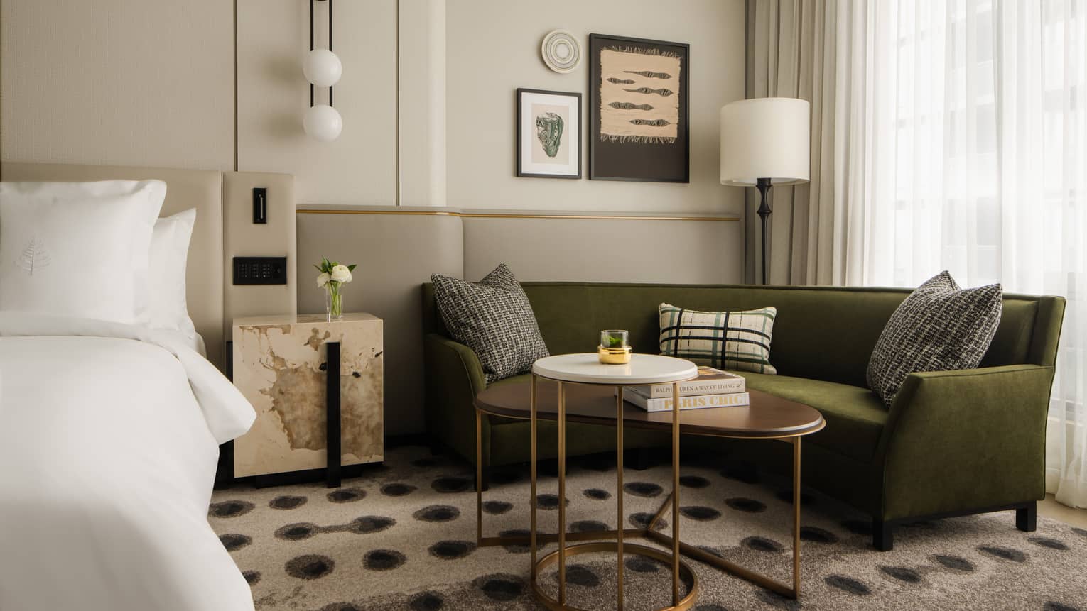 Hotel room lounge area with a green sofa, decorative cushions, modern artwork, adjacent bed and large windows