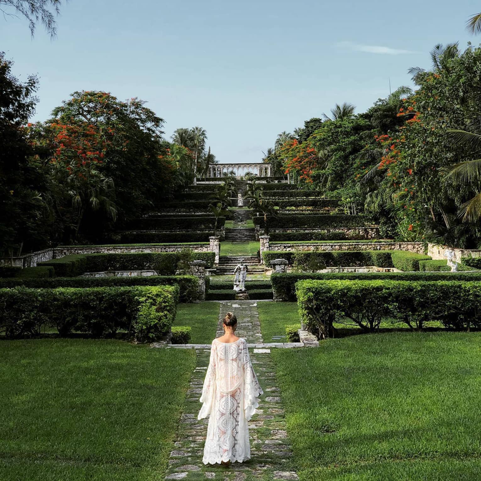 Person in a white flowing dress walking through terraced gardens with stone paths and lush greenery