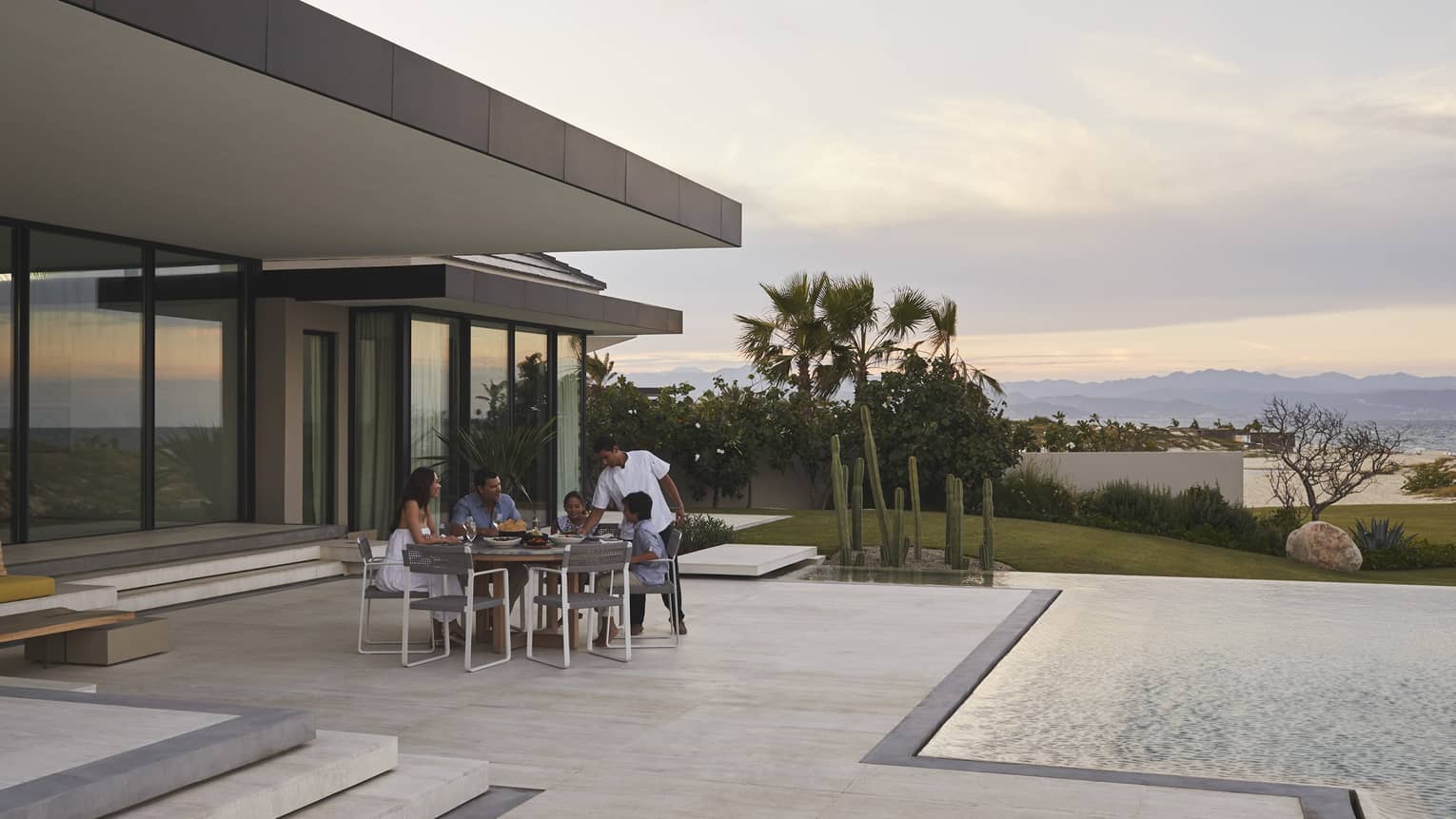 Family dining outdoors on a modern patio by the pool, with a scenic view of mountains and desert plants at sunset, creating a serene and intimate setting.