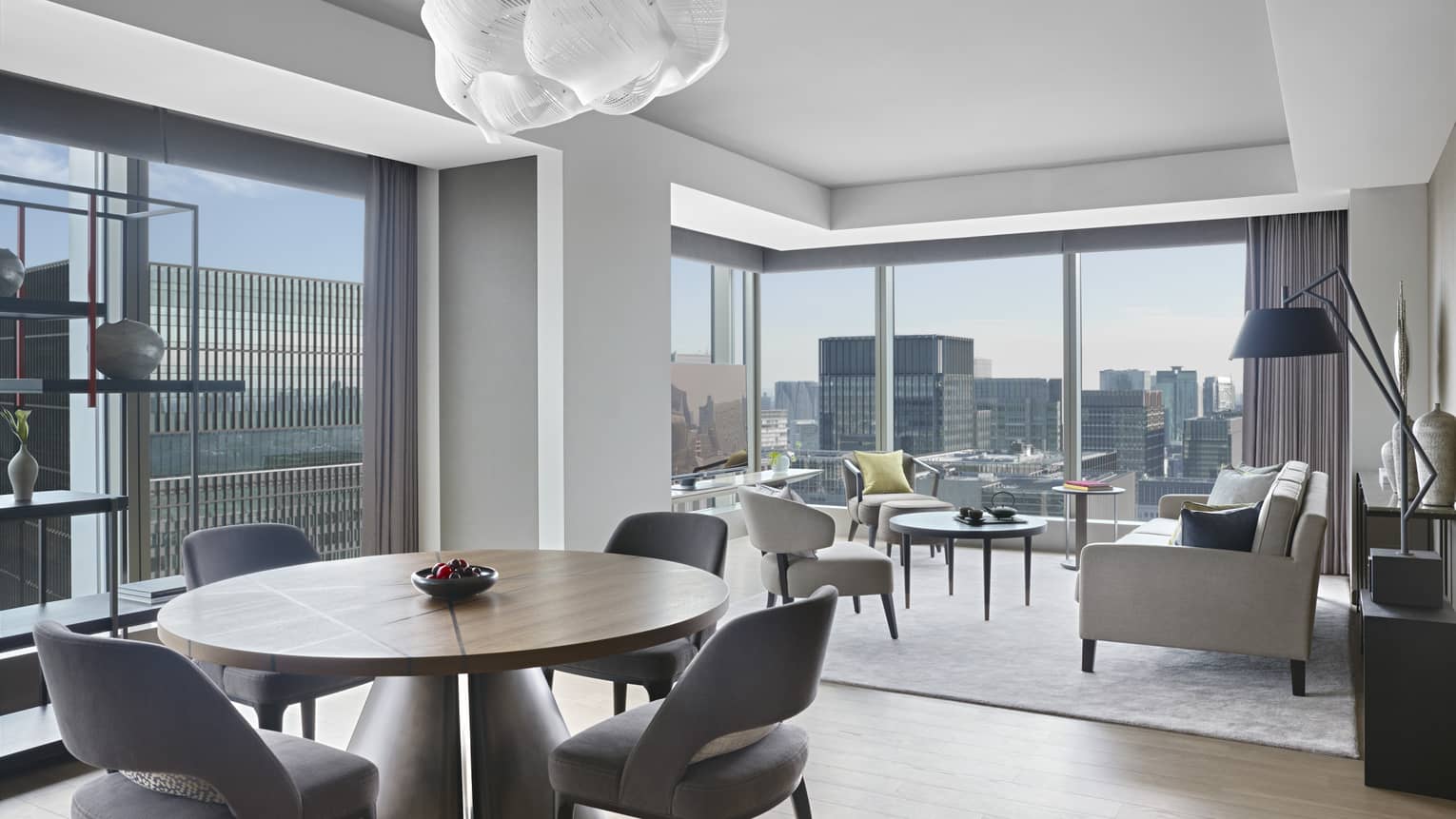 Hotel suite with round dining table with chairs, seating area, modern shelving and floor-to-ceiling windows with panoramic city views