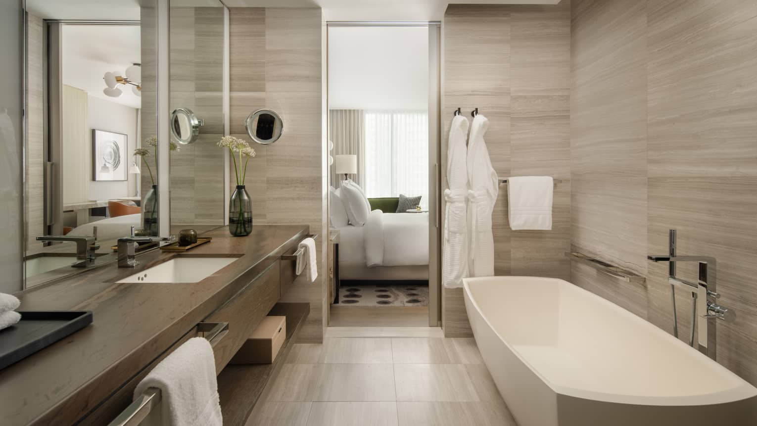 Luxury hotel bathroom with a freestanding tub, spacious vanity, large mirror, and two white bathrobes hanging beside the door