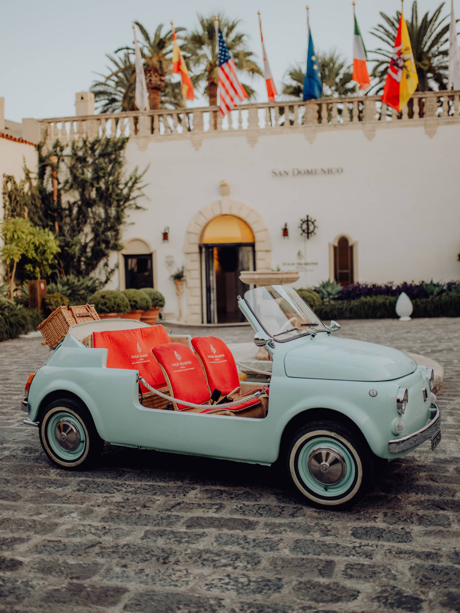 Vintage teal convertible Fiat 500 with bright coral seats parked on a cobblestone driveway in front of San Domenico Palace.
