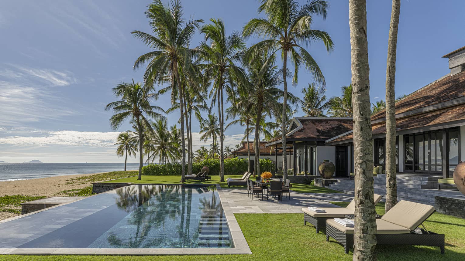 Three Bedroom Beachfront Pool Villa pool area with dining table, lounge chairs, palm trees and blue skies