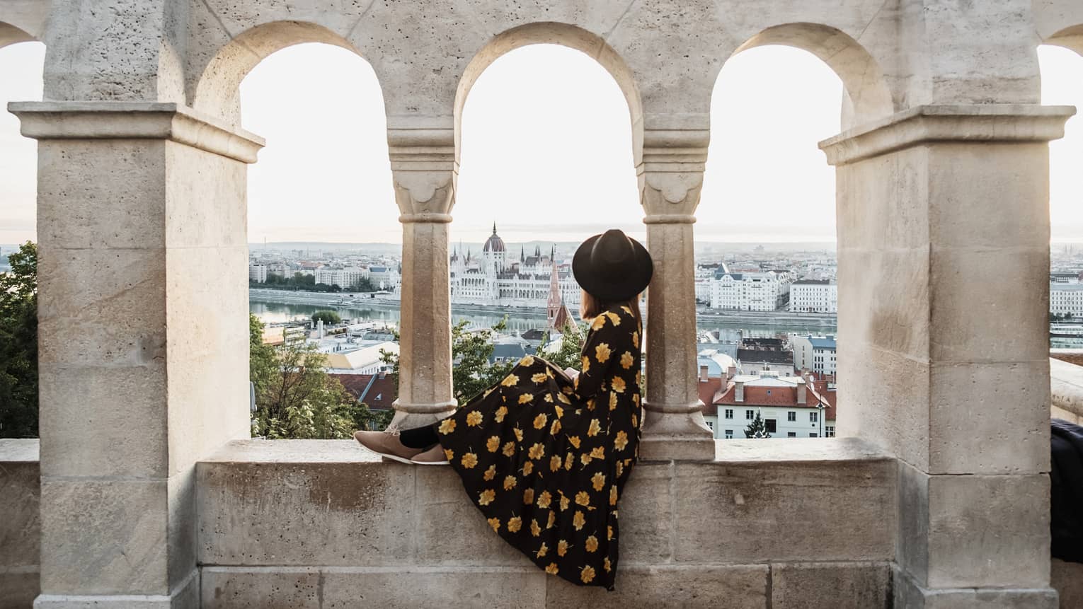 A guest in a floral dress and hat sitting between stone arches, gazing at a distant cityscape and river.