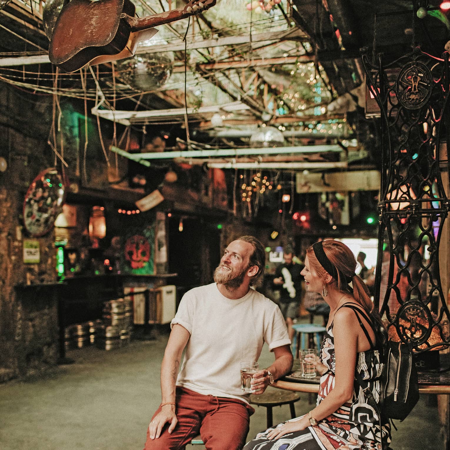 A smiling couple holding drinks, surrounded by myriad disparate objects and scrap parts on the walls and hanging overhead.