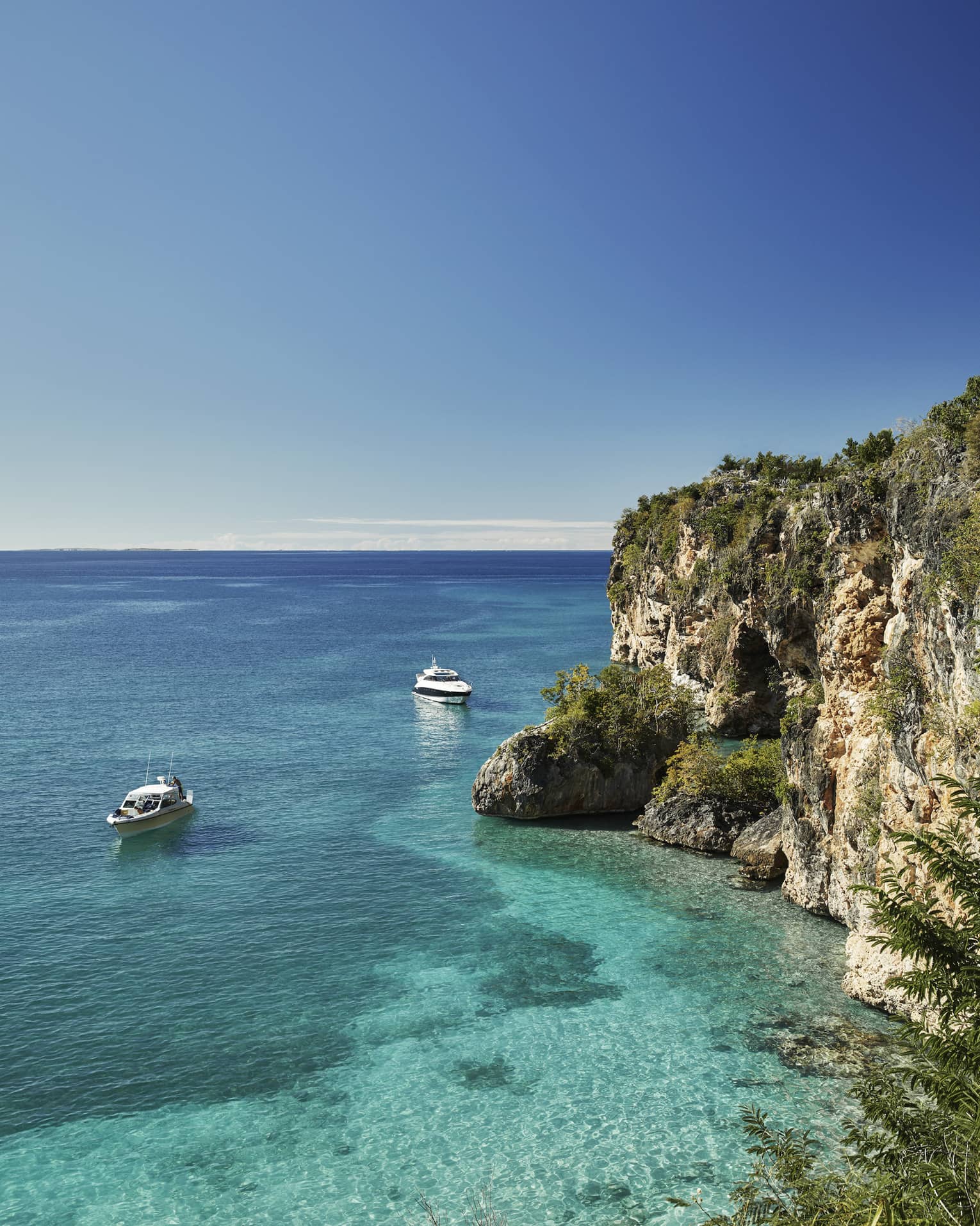Long view of two cabin boats floating in the crystal-clear ocean beside towering rocky cliffs, a pale blue sky above.