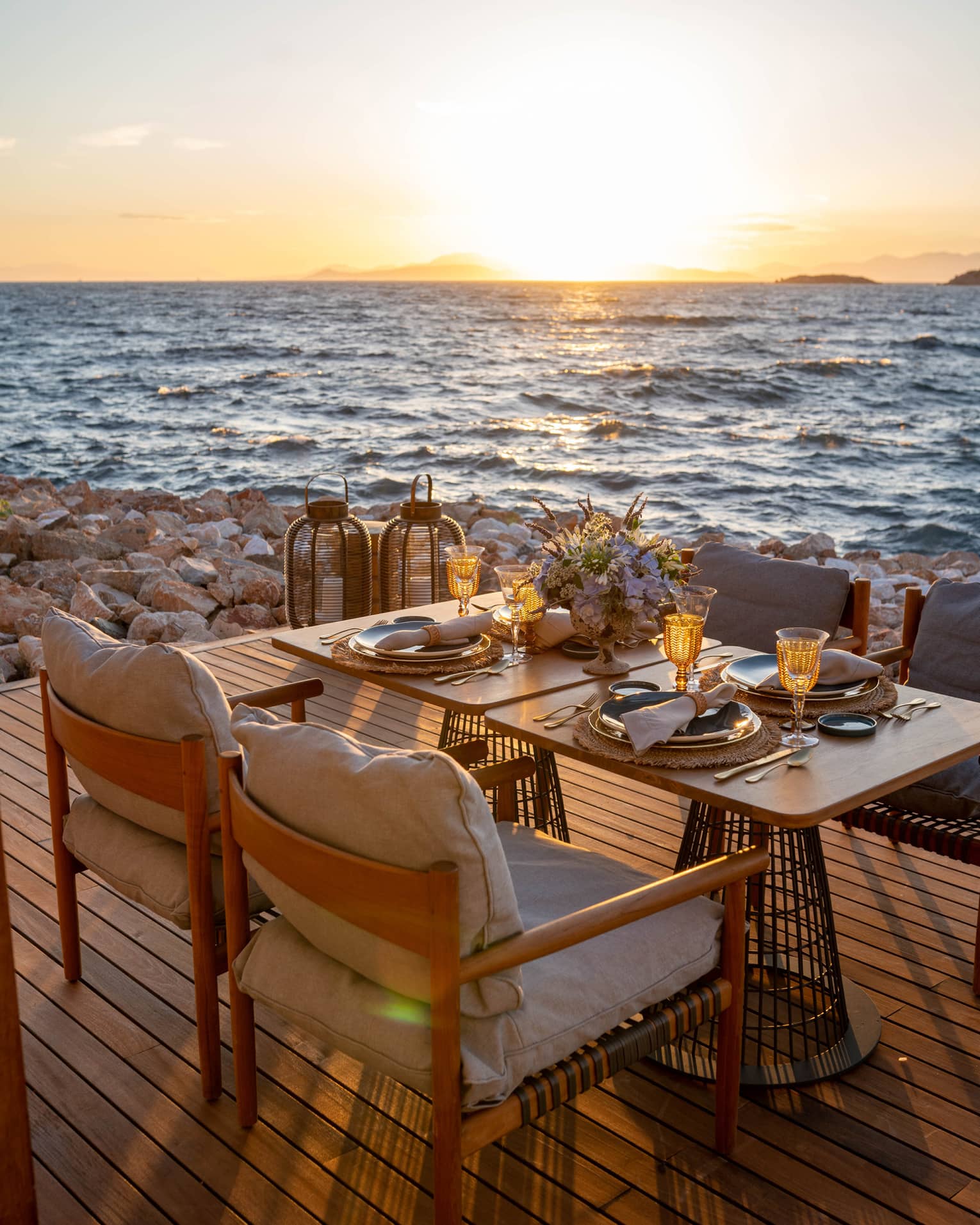 A table laid with sparkling glasses and a bouquet of flowers set on a deck on the rocky shore, the sunlit waves metres away.