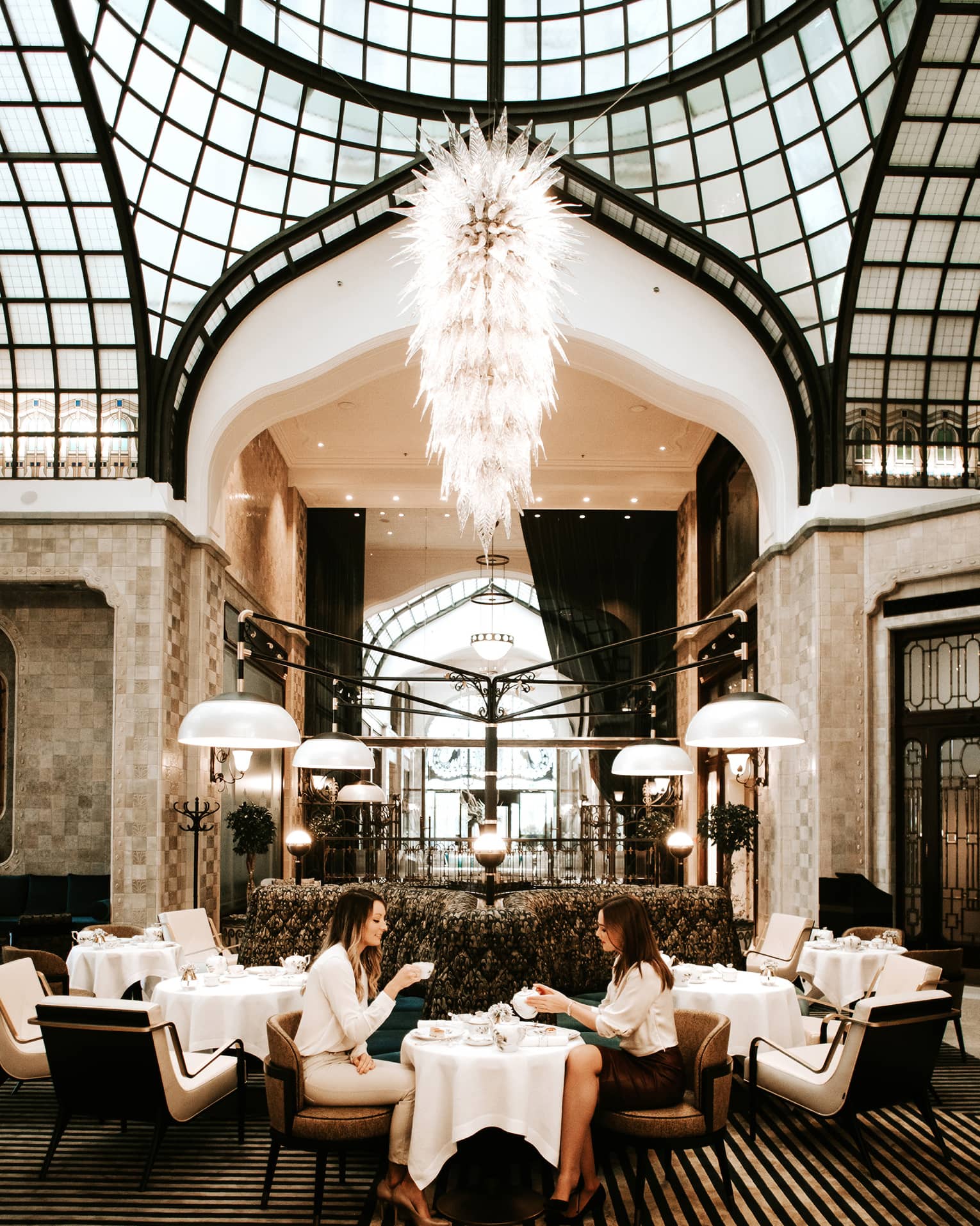 Elegant dining area with a grand chandelier, high arched glass ceiling and modern seating, where two people are enjoying tea at a small table