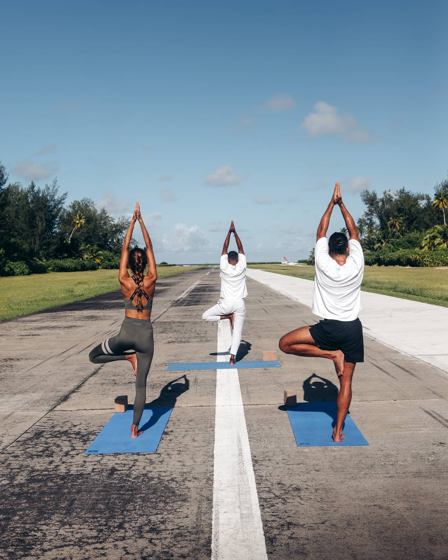 Rear view of three people practicing yoga in tree pose on an airport runway framed by lush greenery, under a clear blue sky.