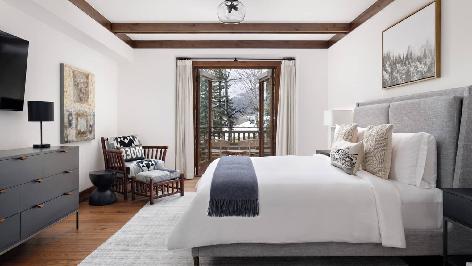 Luxury resort bedroom with a grey upholstered king bed, wall artwork, wooden armchair, dresser with black lamp, and open doors to a snowy forest-view balcony