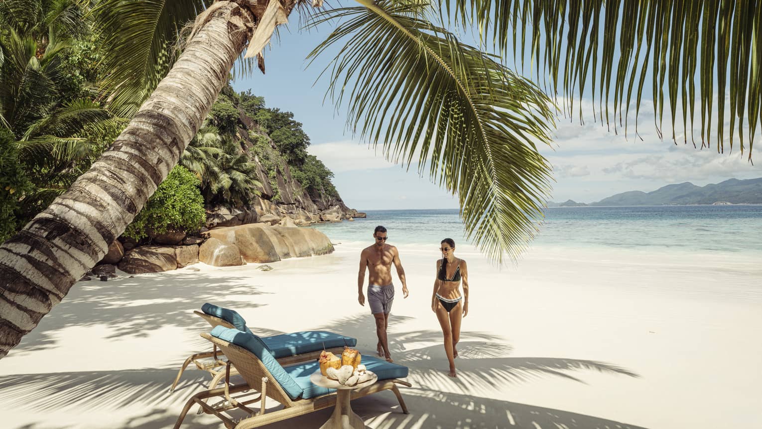 Couple walking side by side towards two lounge chairs under a palm tree on a white-sand beach