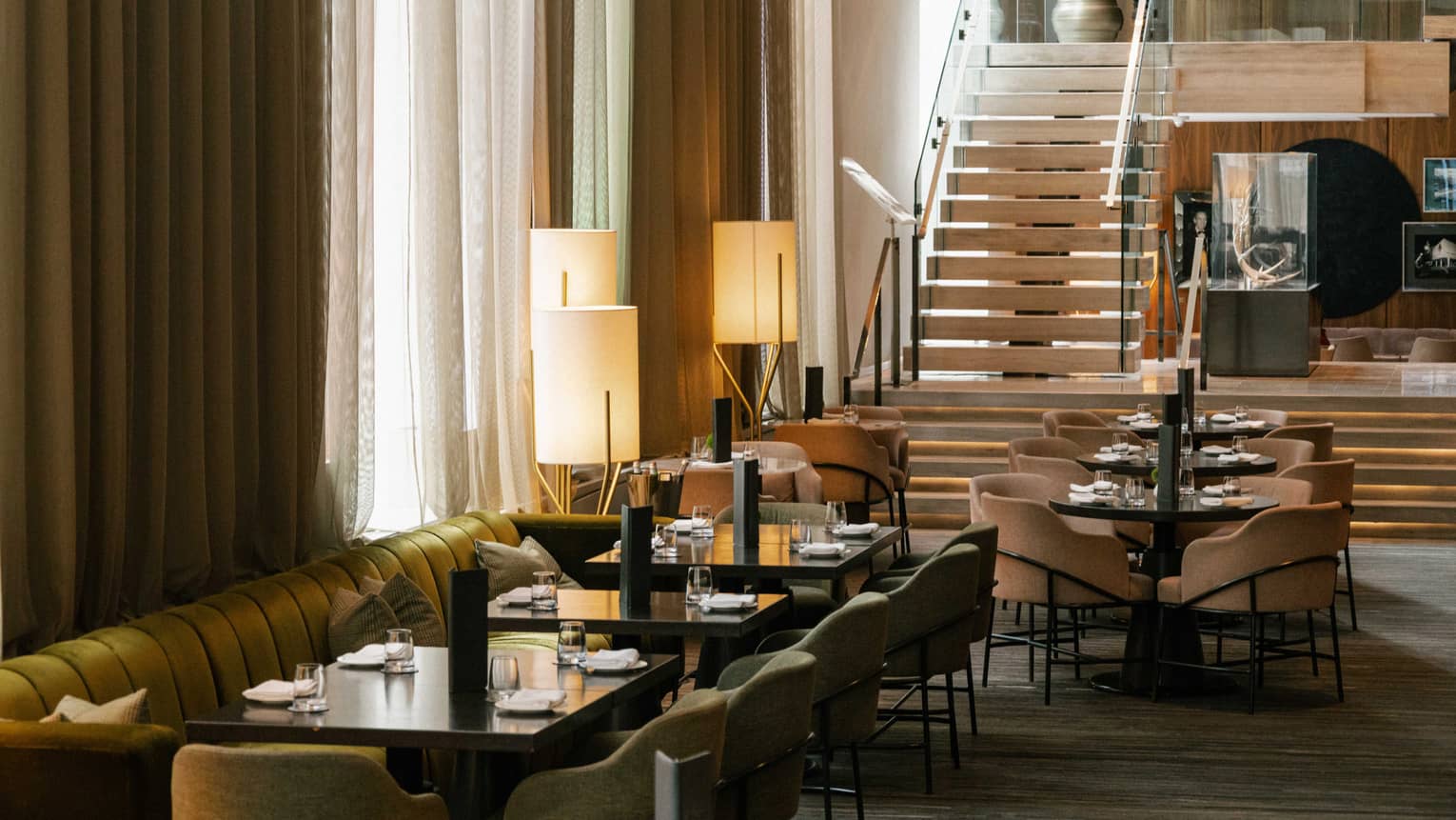 A dining room with a velvet upholstered booth, wooden tables and cushioned chairs with modern light fixtures and large windows