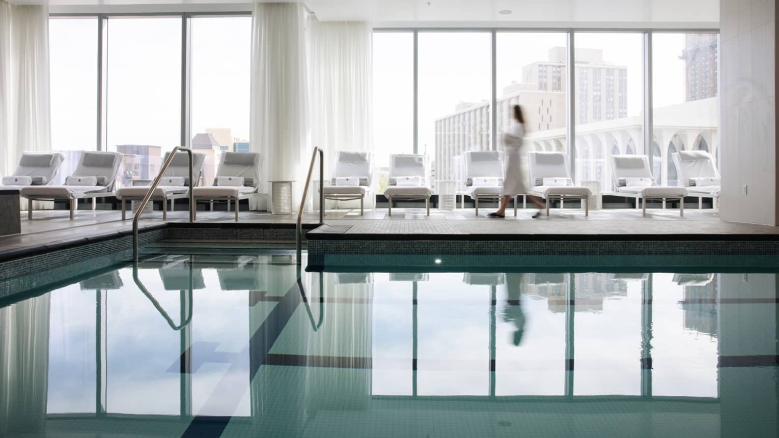 A woman in a spa robe walking along lounge chairs by an indoor pool.