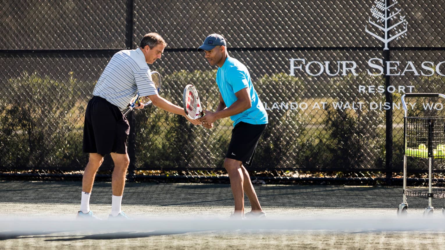 Tennis pro helps man adjust his racket on tennis court by Four Seasons Resort logo on fence