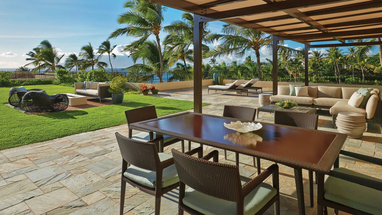 Covered patio with dining table, lounge seating and ocean view surrounded by palm trees and greenery