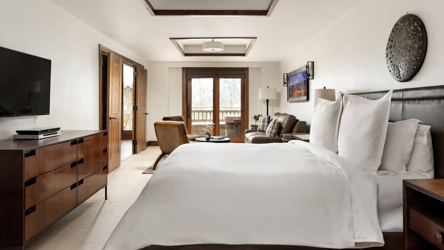 Bedroom with a king bed, wall-mounted TV, dresser and cozy seating area by wooden-framed doors leading to a balcony
