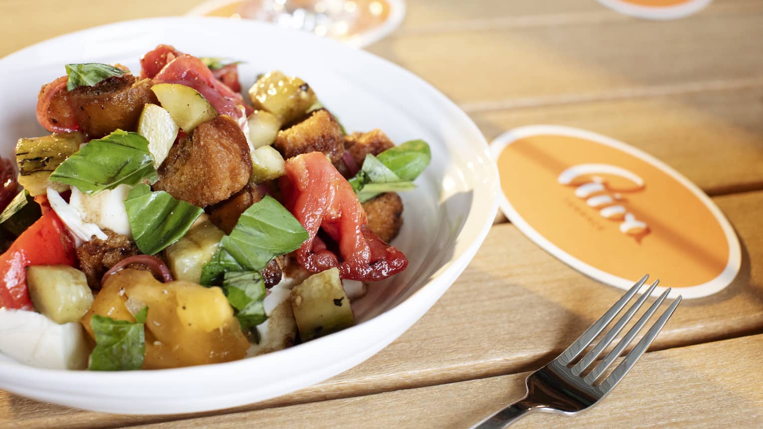 Hearty salad in a white bowl next to a silver fork and an orange coaster with the name "Riva" on it, all on a wooden table