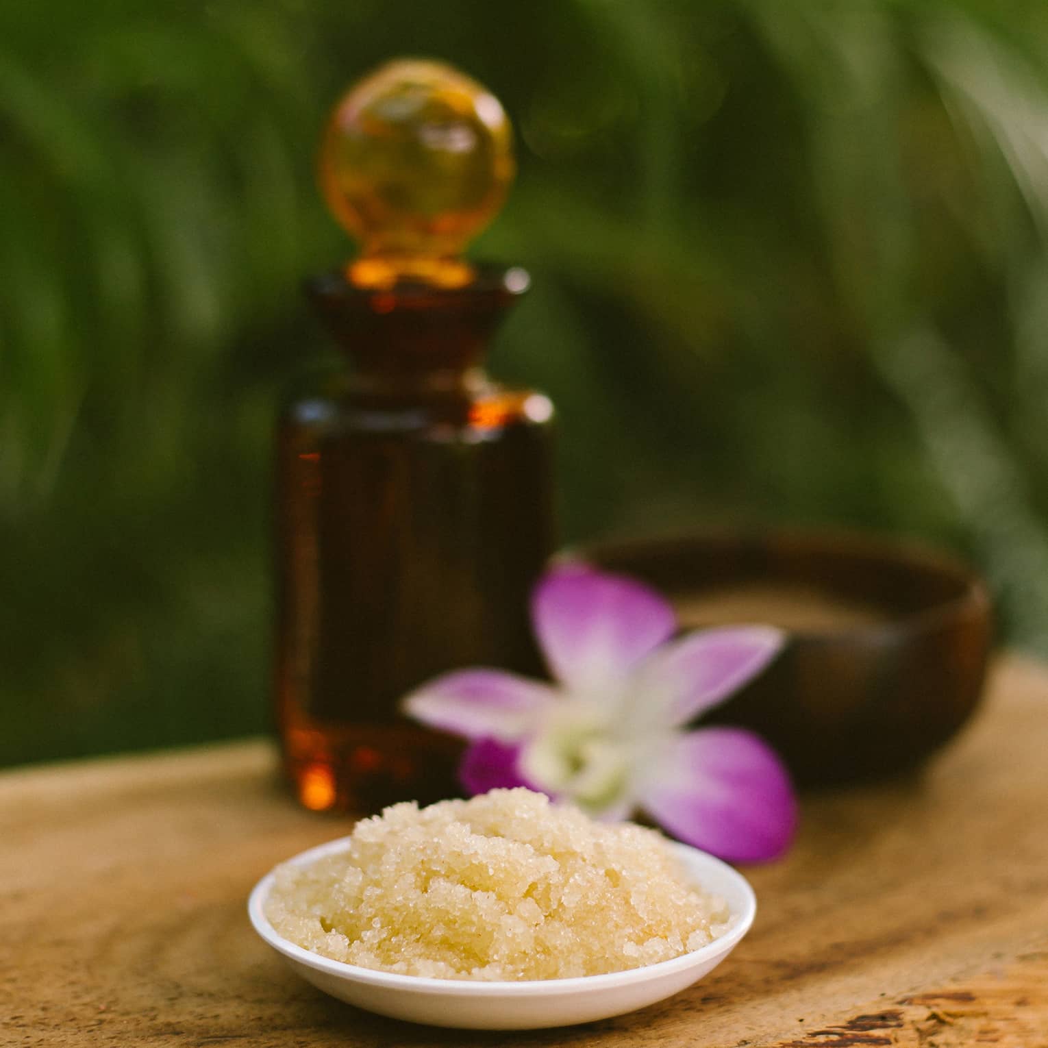 Bowl with salt scrub next to purple tropical flower and glass oil bottle on rustic table