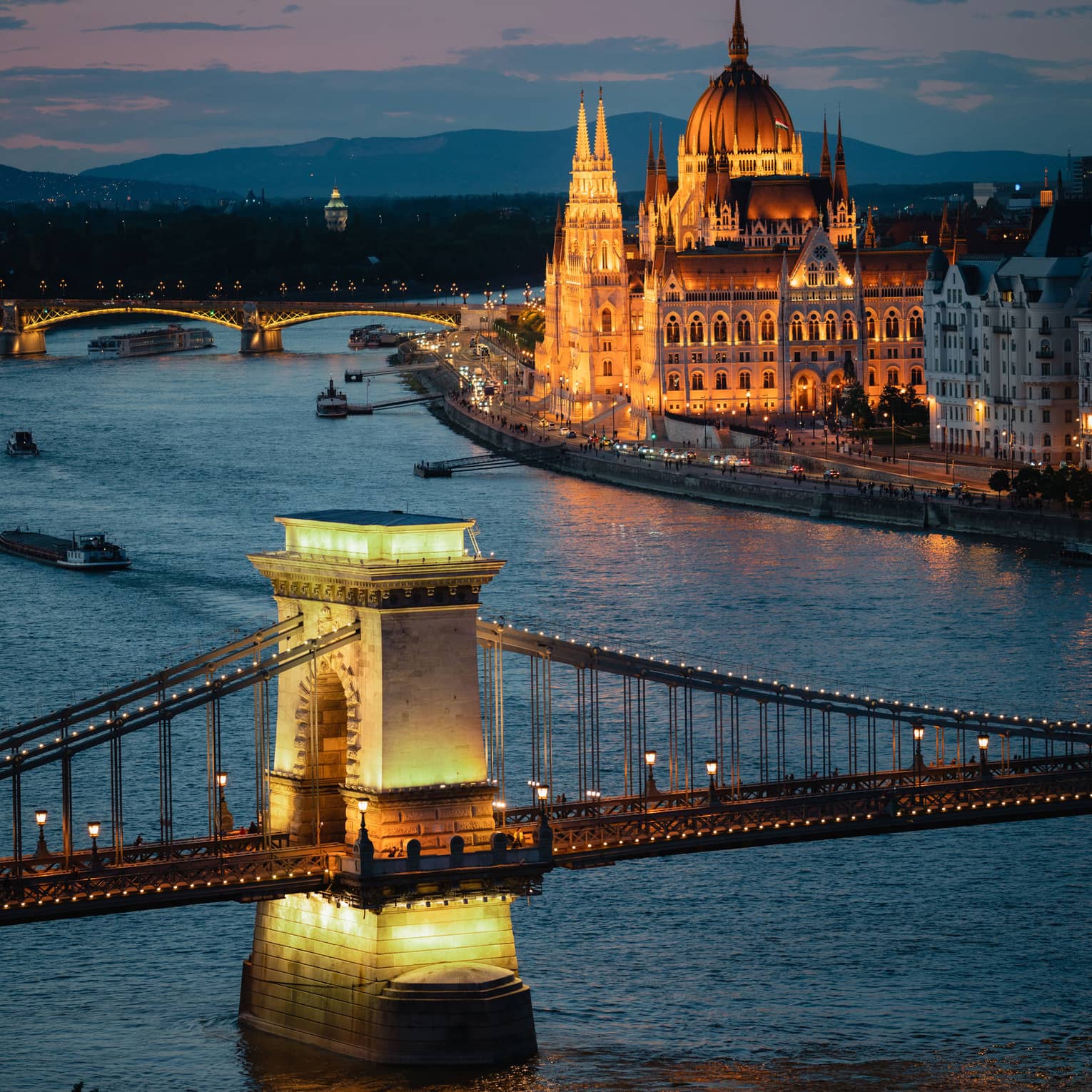 Illuminated bridge spanning a river at dusk, with a grand, lit-up building and boats along the waterfront.