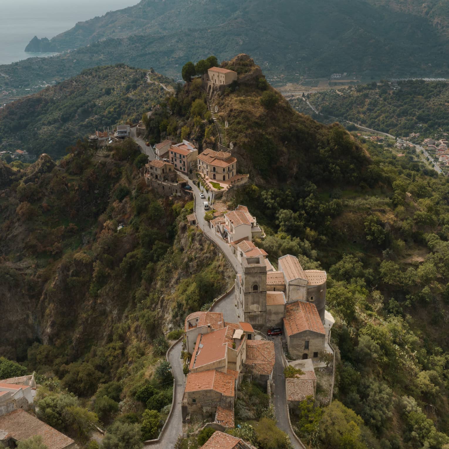 Aerial view of a historic hilltop village with winding roads, stone buildings and terracotta roofs, nestled in a lush green mountain landscape.