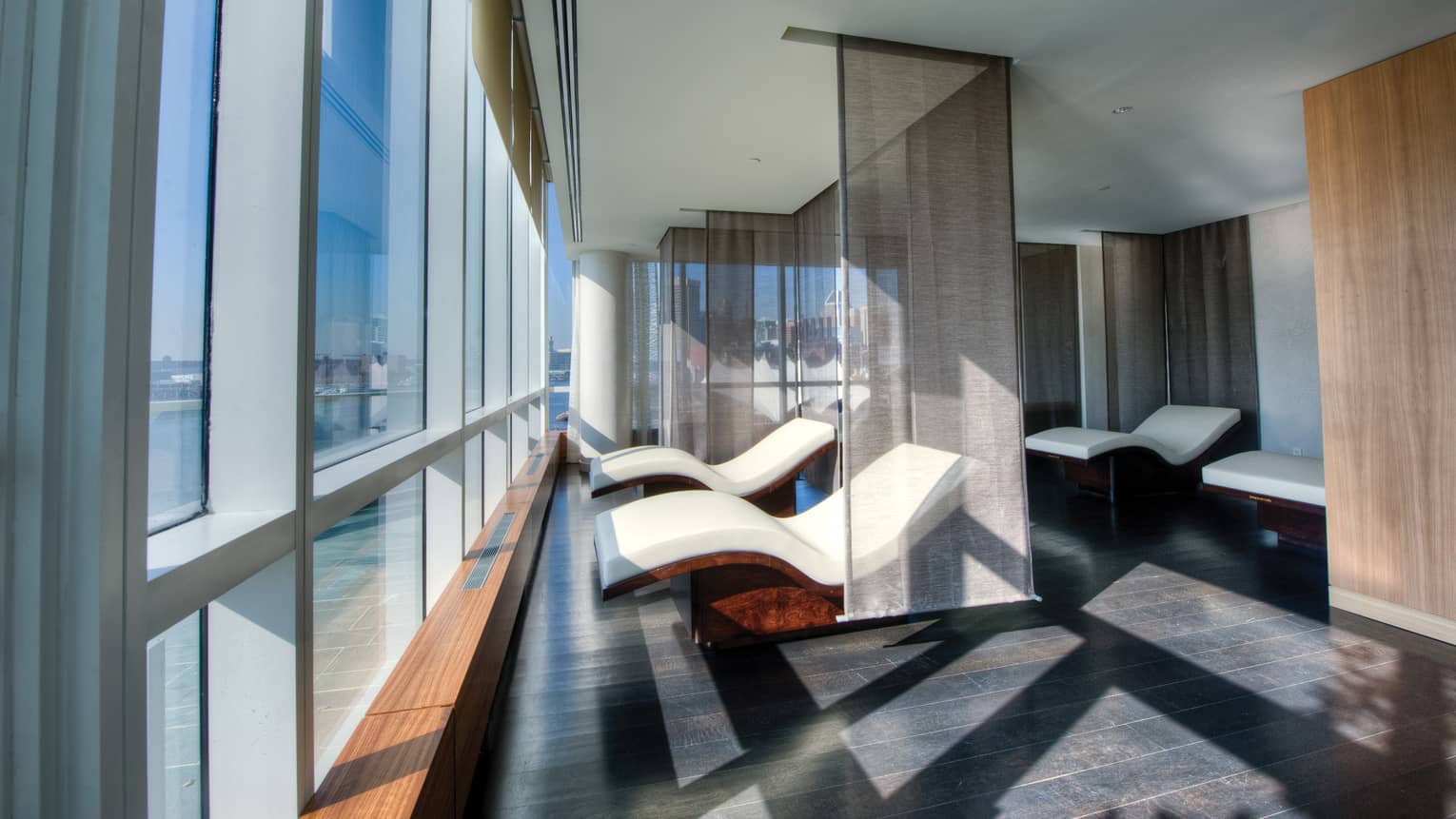 Two curved white spa chairs divided by sheer panel curtains in front of floor-to-ceiling windows