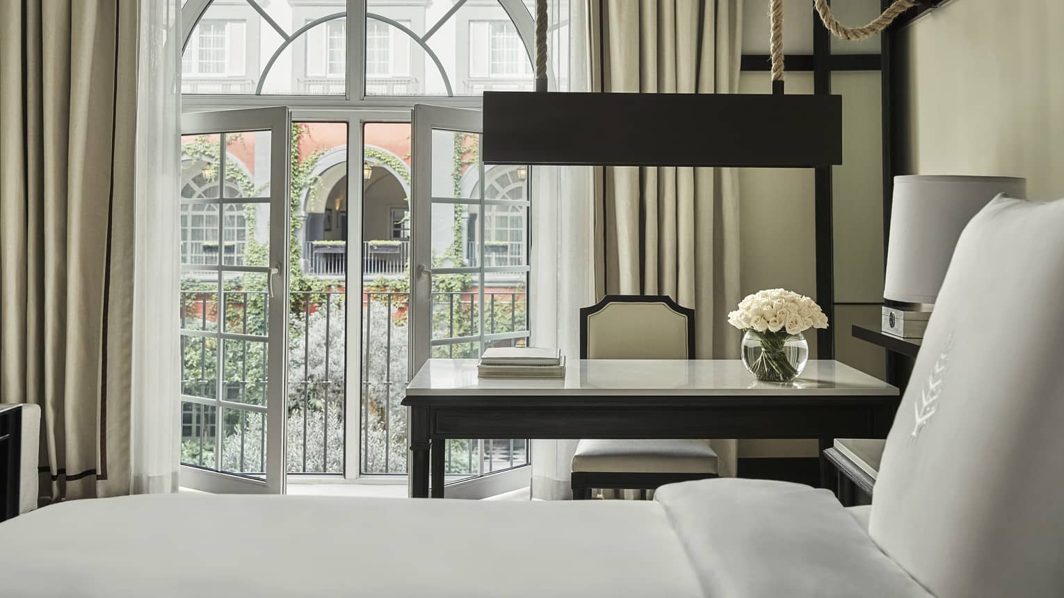 Elegant hotel room with a large arched window, a desk with flowers, and an inviting view of a garden courtyard framed by ivy-covered walls.