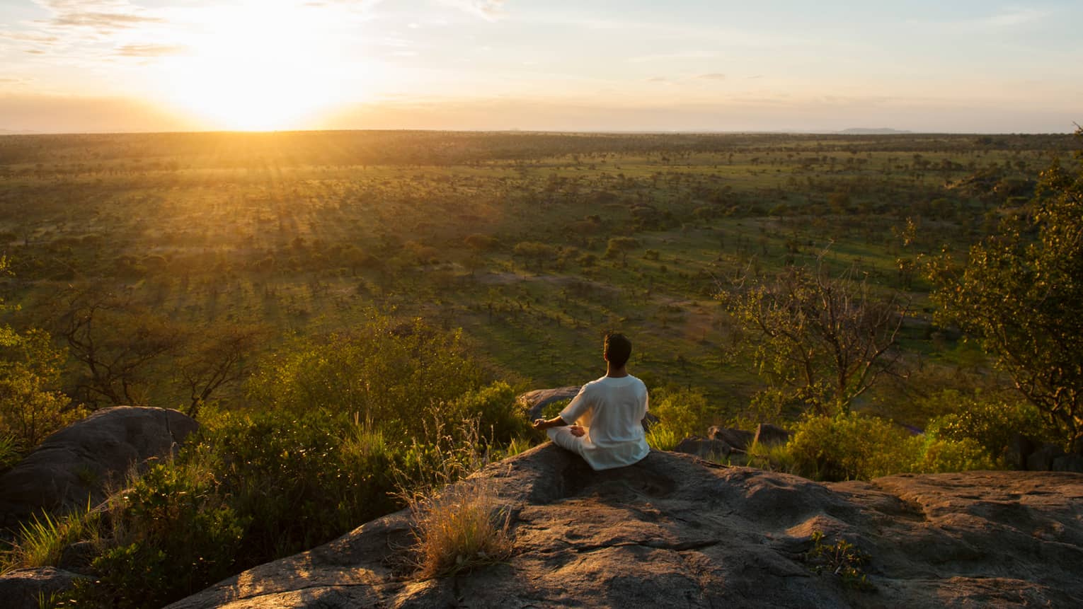 Woman sits crossed-legged, meditates on cliff overlooking Serengeti sunset