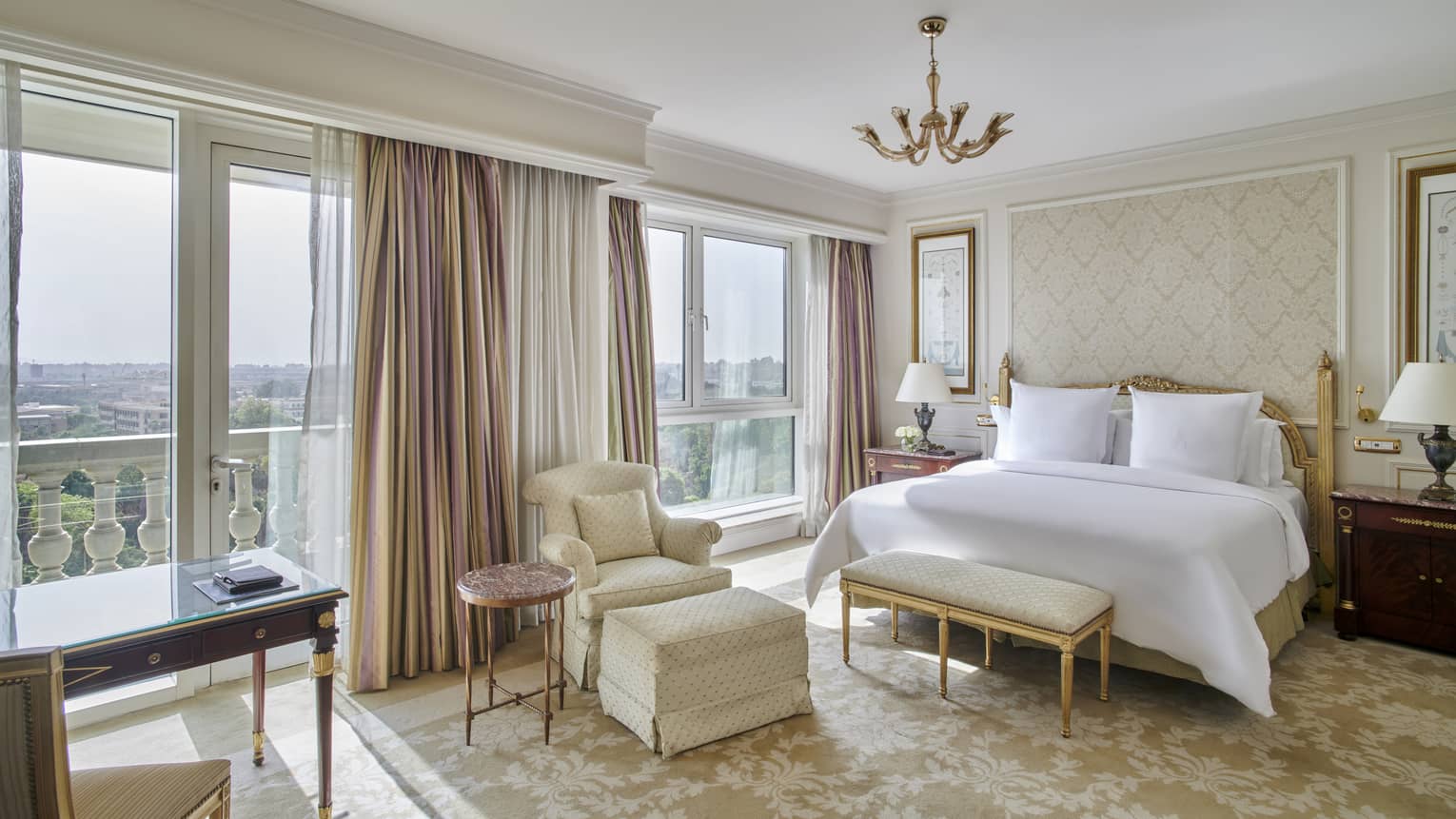 Guest room with king bed and white linens, foot bench, arm chair, floor-to-ceiling windows with city views