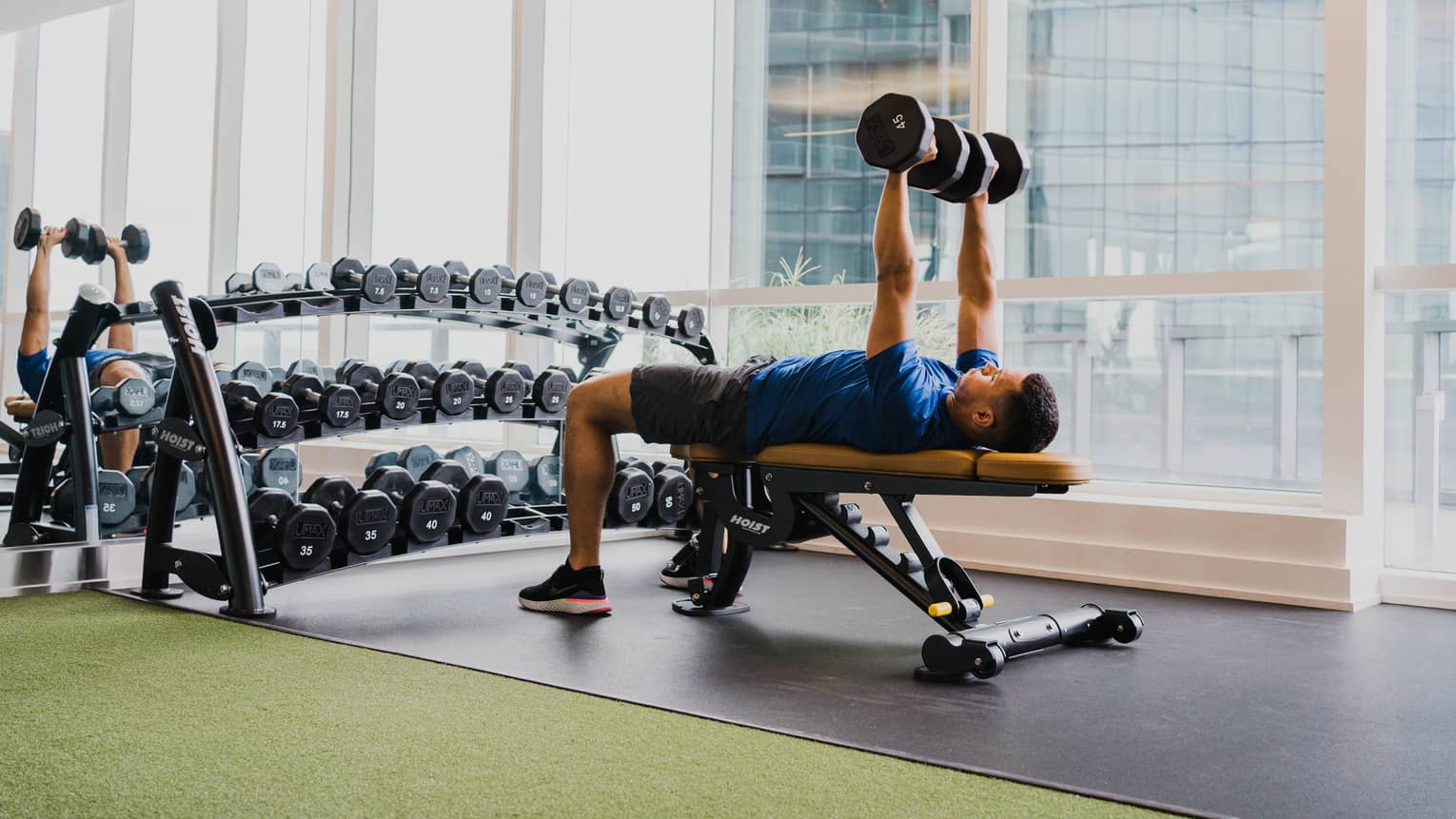 A man working out with free weights in a clean fitness room with large windows.