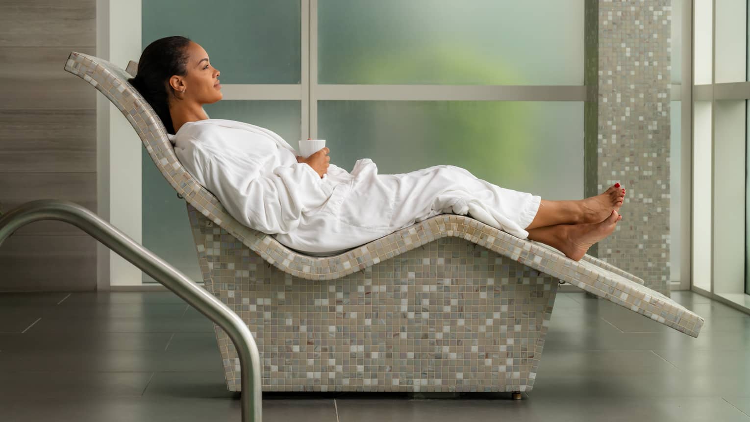Guest wearing a robe and laying in a Spa chair in a room with floor-to-ceiling windows.