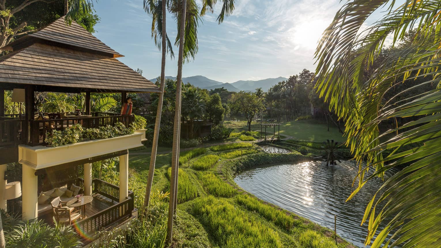 Woman stands on second floor balcony, looks beyond palms to green grass, river below