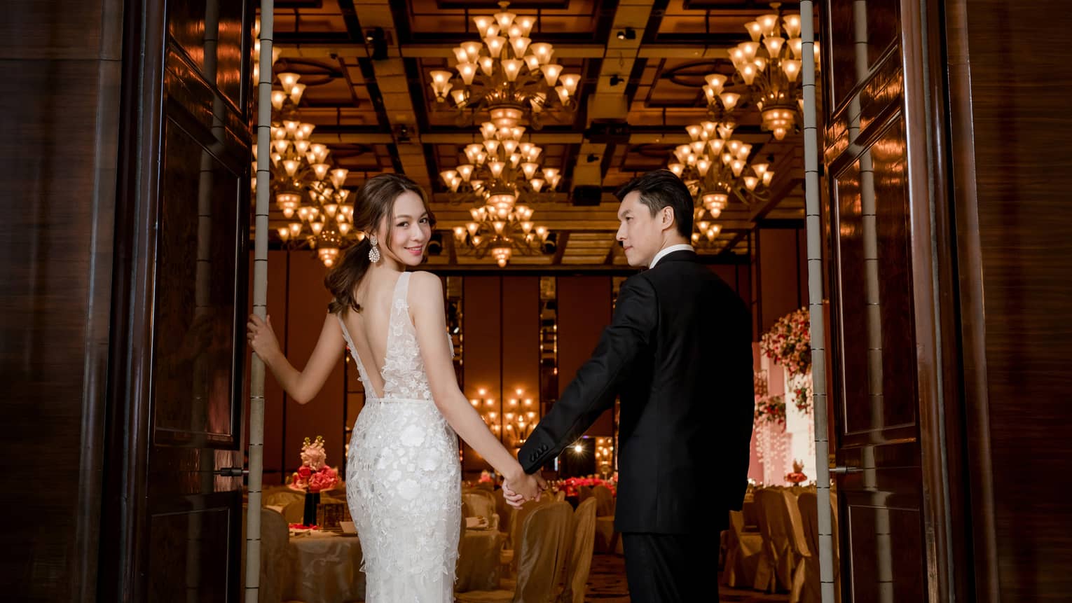A bride and groom enter through the doors of a grand reception room decorated with banquet tables and ornate chandeliers