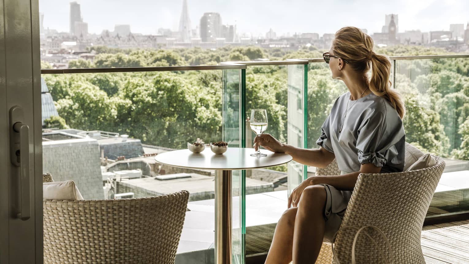 A guest takes in the city skyline while comfortably relaxing in a wicker chair and enjoying a drink on a balcony.