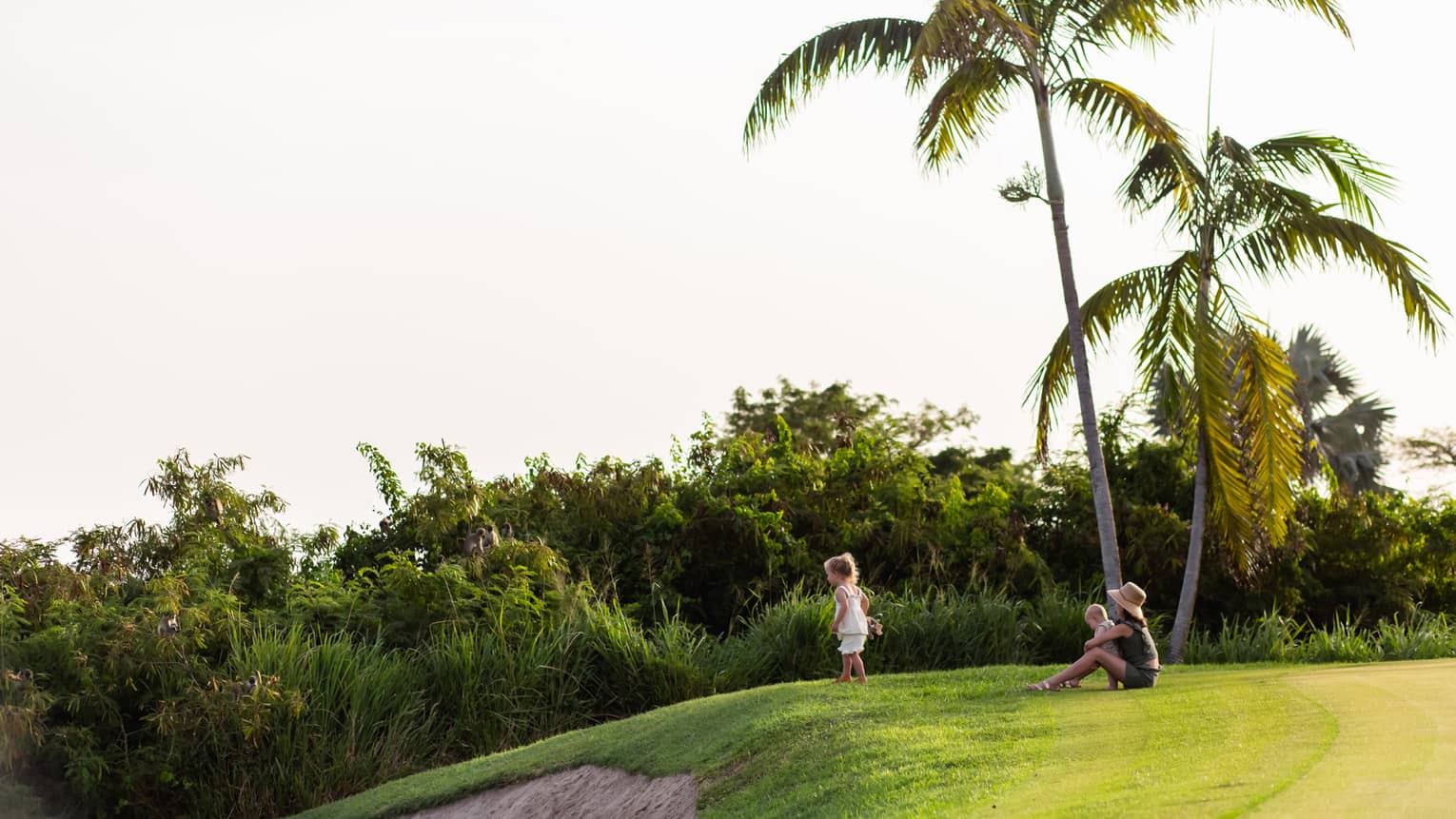 An adult and two small children sit on the edge of a hillside golf course overlooking palm trees and jungle