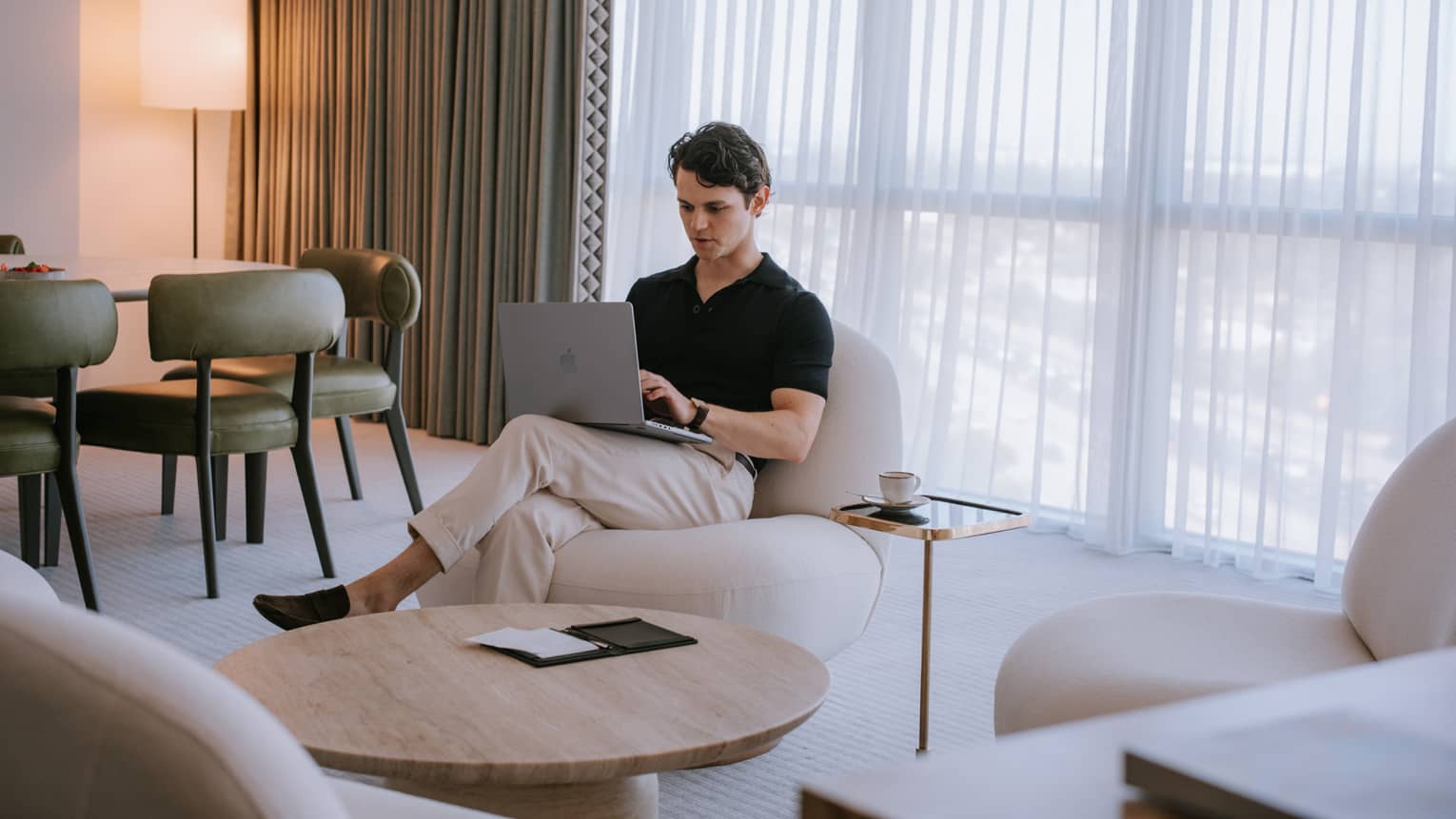 Person sitting in a living room with floor-to-ceiling windows and typing on a laptop.