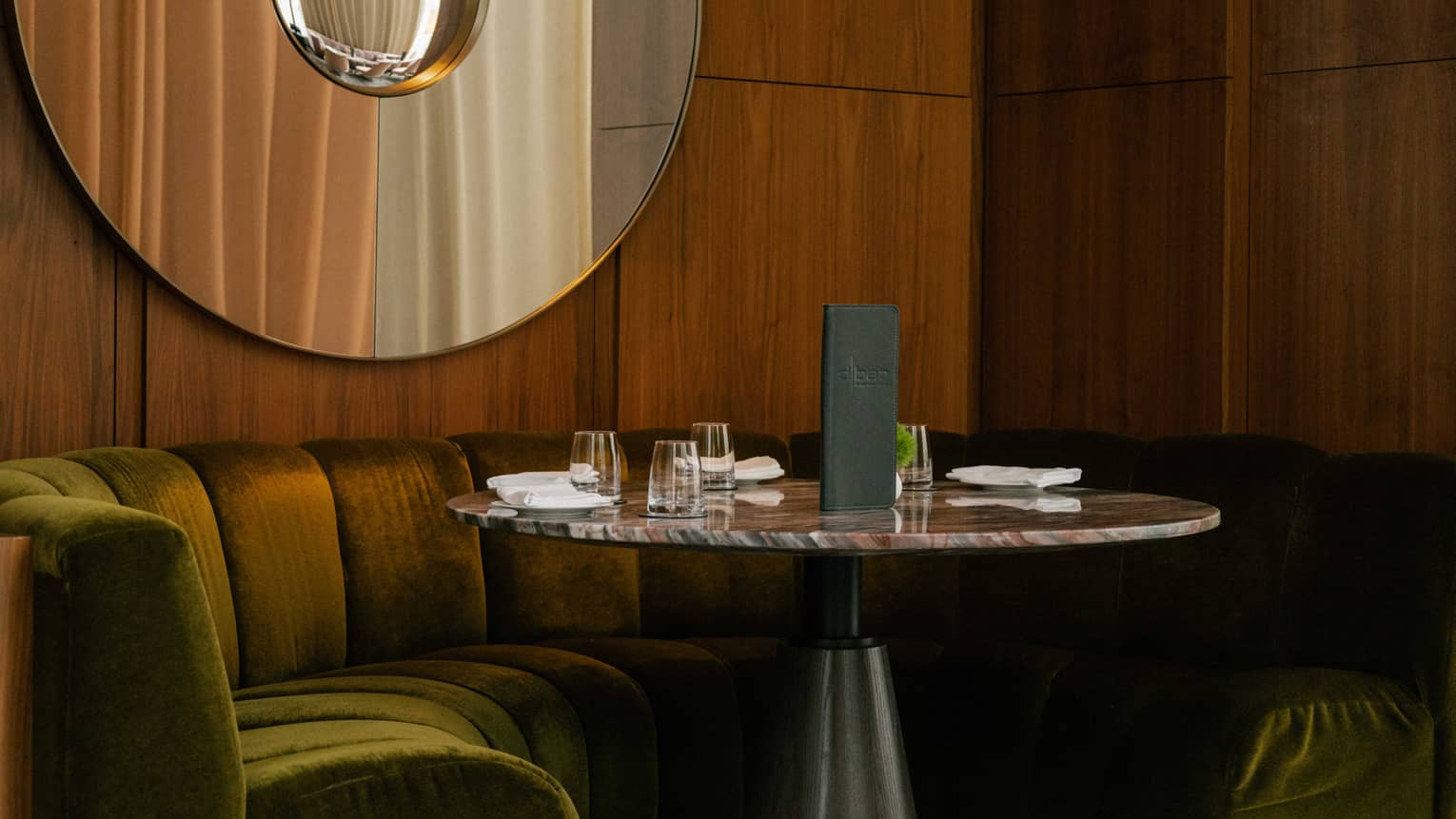 Close up view of a corner booth with green velvet seats and a round marble table below a modern art piece
