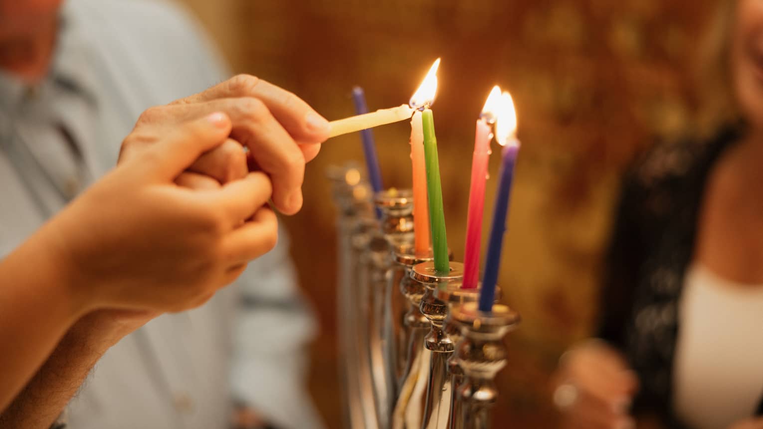 Two hands light multi-coloured candles on a menorah
