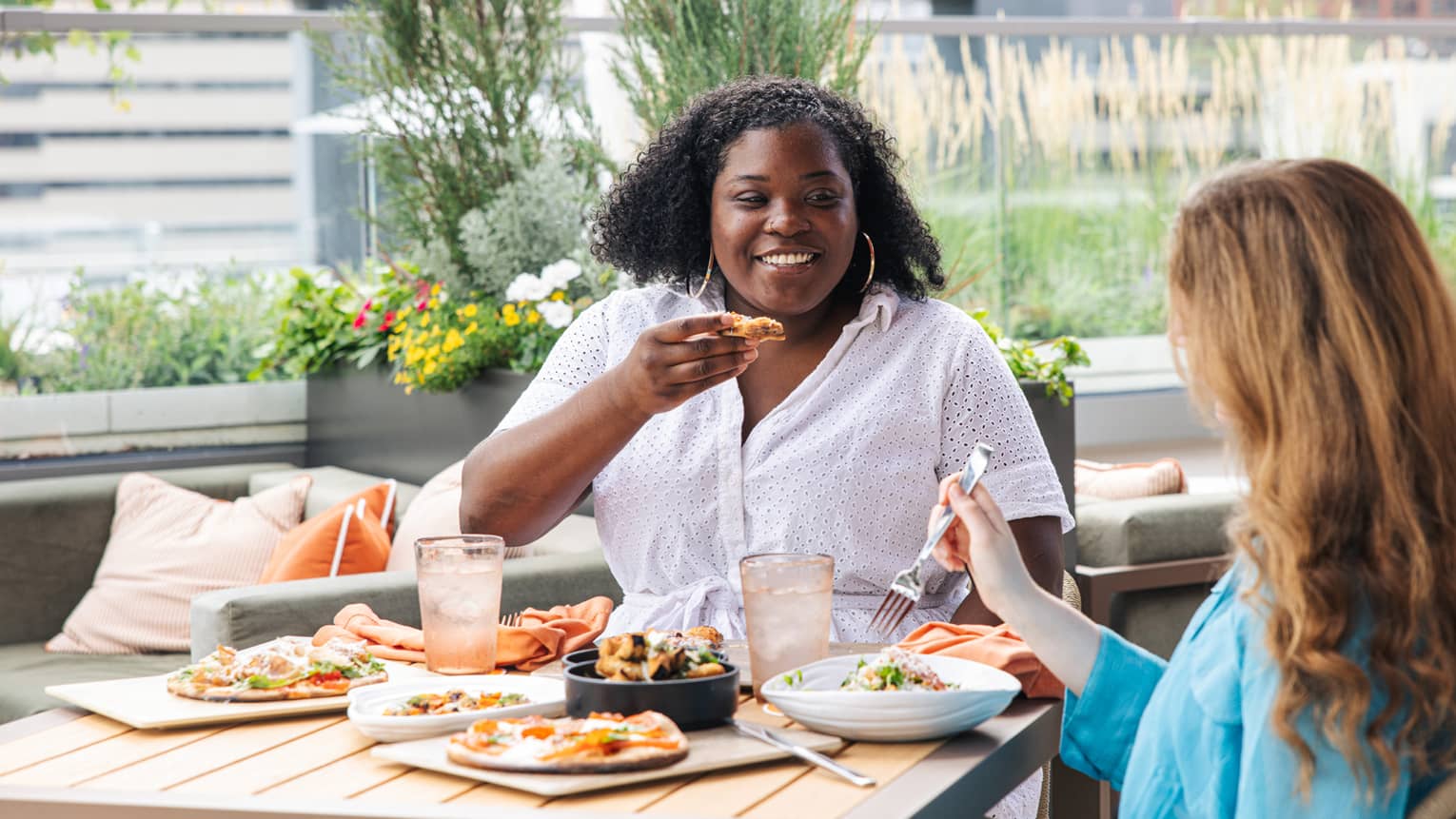 Two guests dine at an outdoor terrace surrounded by florals and greenery with a city skyline behind