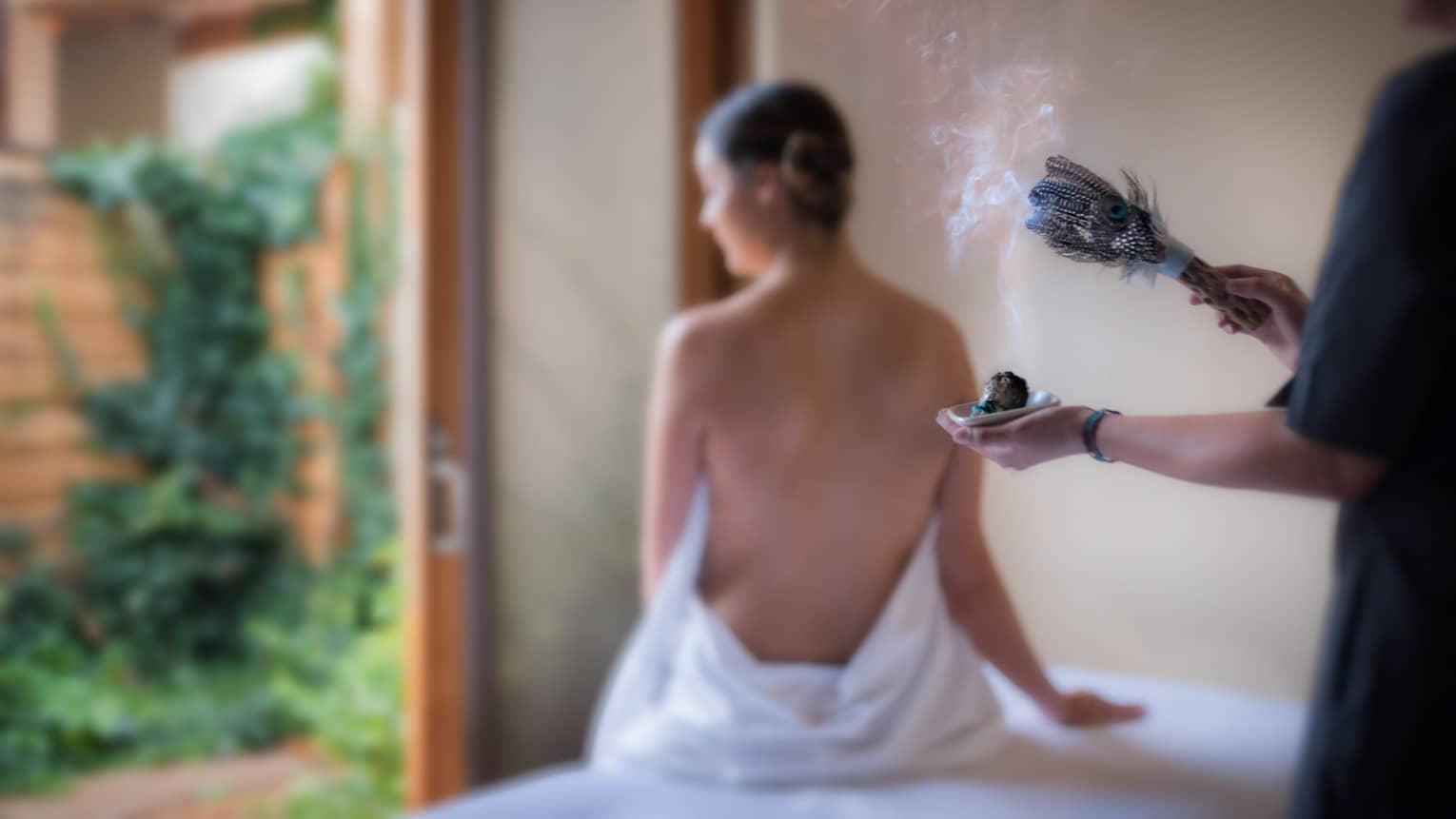 Smoke rises from sage brush with feathers as spa staff performs smudging ceremony for woman
