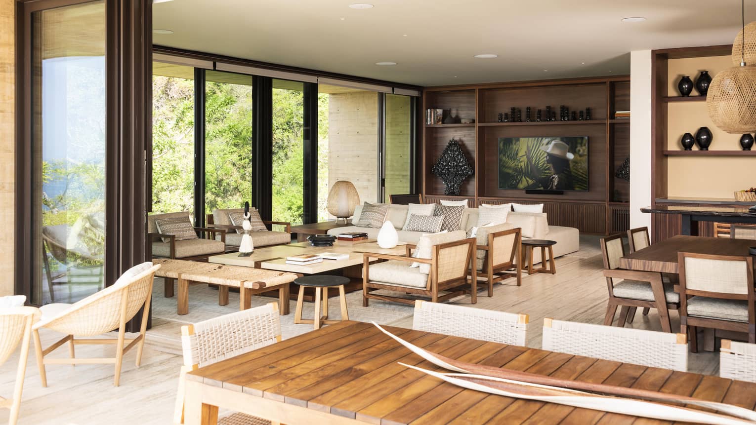 Spacious living area with floor-to-ceiling glass doors, wooden furniture and neutral decor. The room opens to an outdoor view with greenery, blending indoor and outdoor spaces.