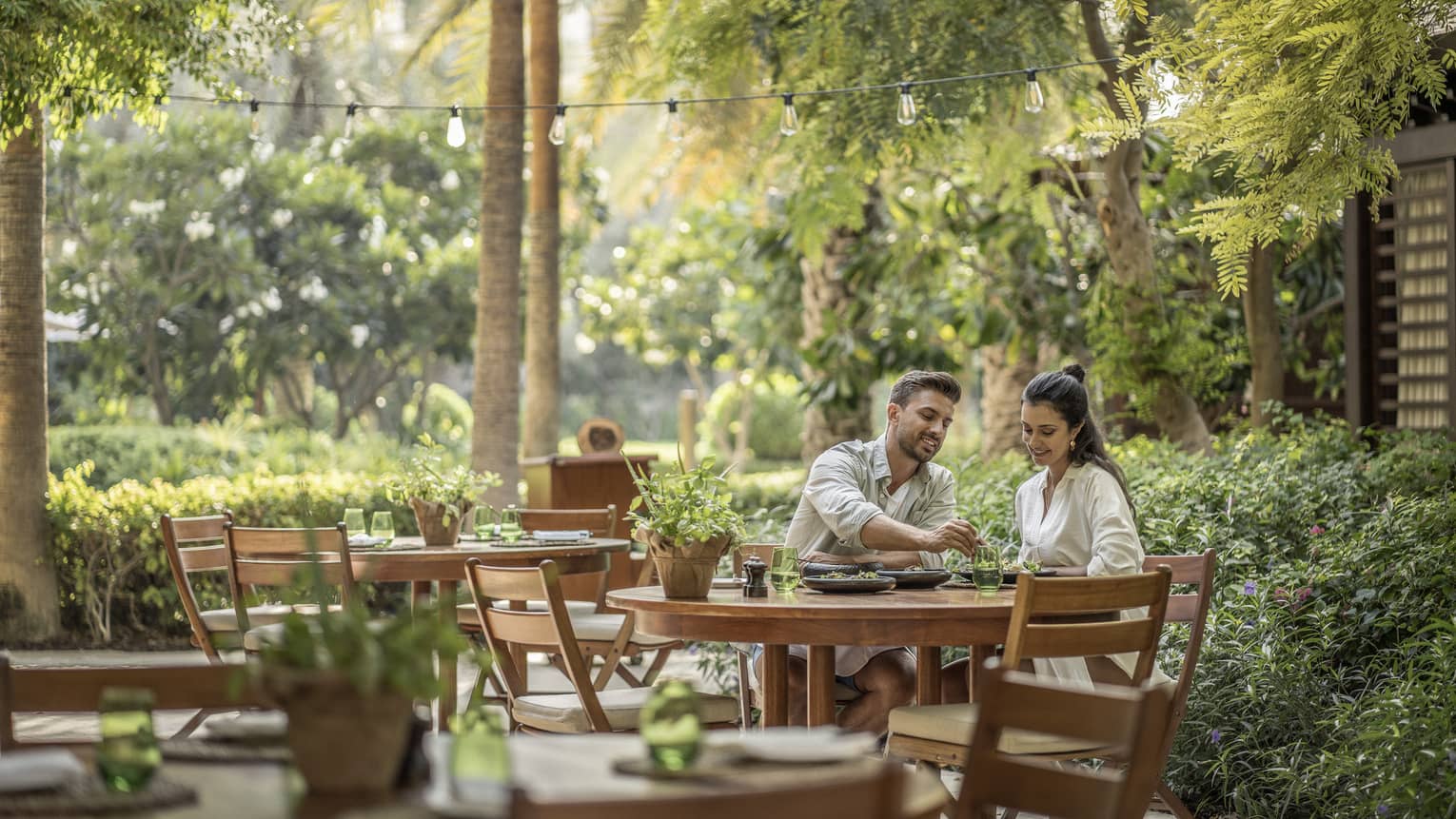 A couple sits at a wooden table on outdoor restaurant terrace, surrounded by greenery