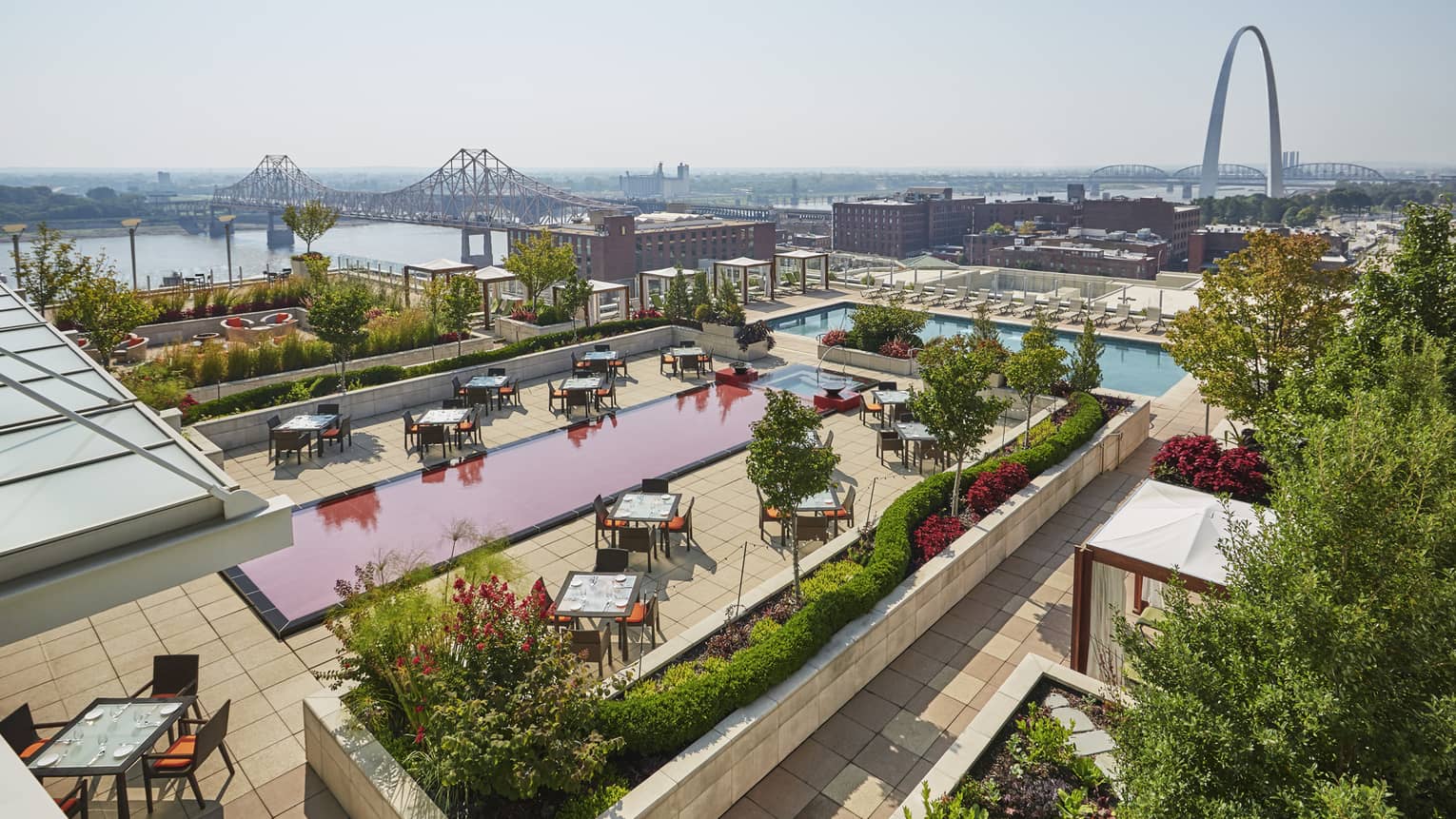 Hotel rooftop terrace with dining tables and a red and blue swimming pool, overlooking a cityscape, river and the Gateway Arch in the distance