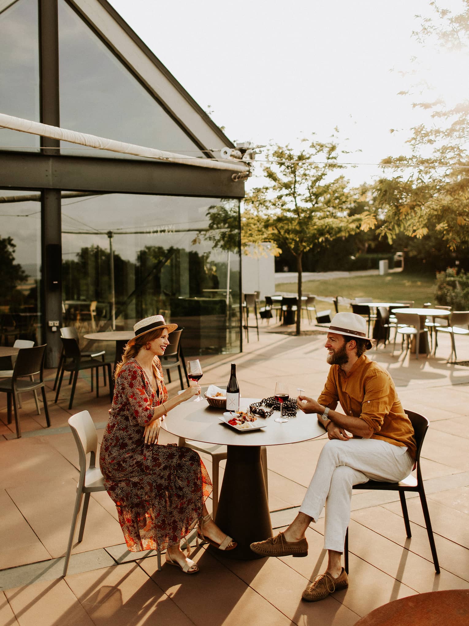 Couple wearing hats sit at outdoor table with plate of food, drinking red wine