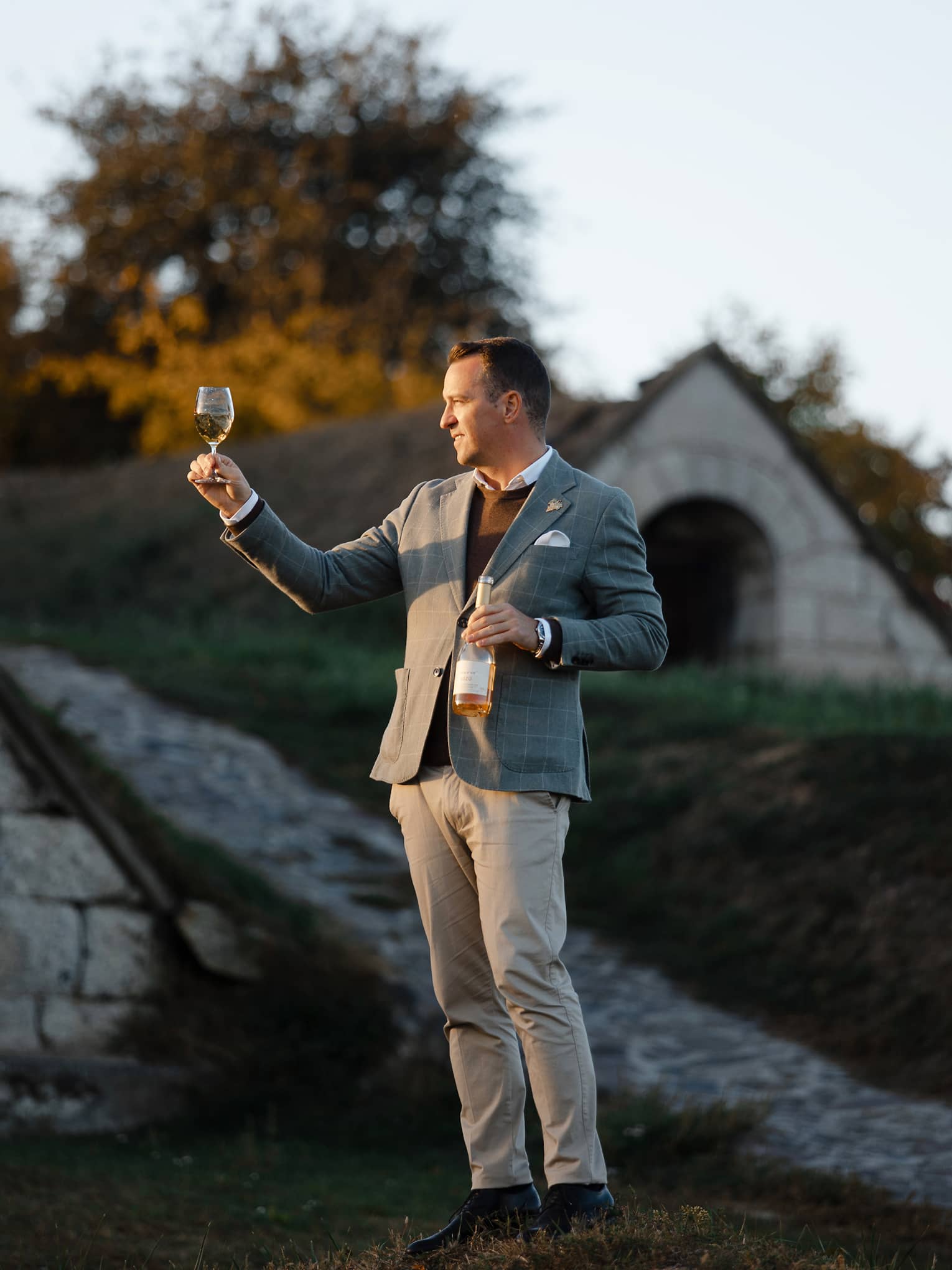 Person in a suit holding a wine bottle and inspecting a glass of white wine outdoors