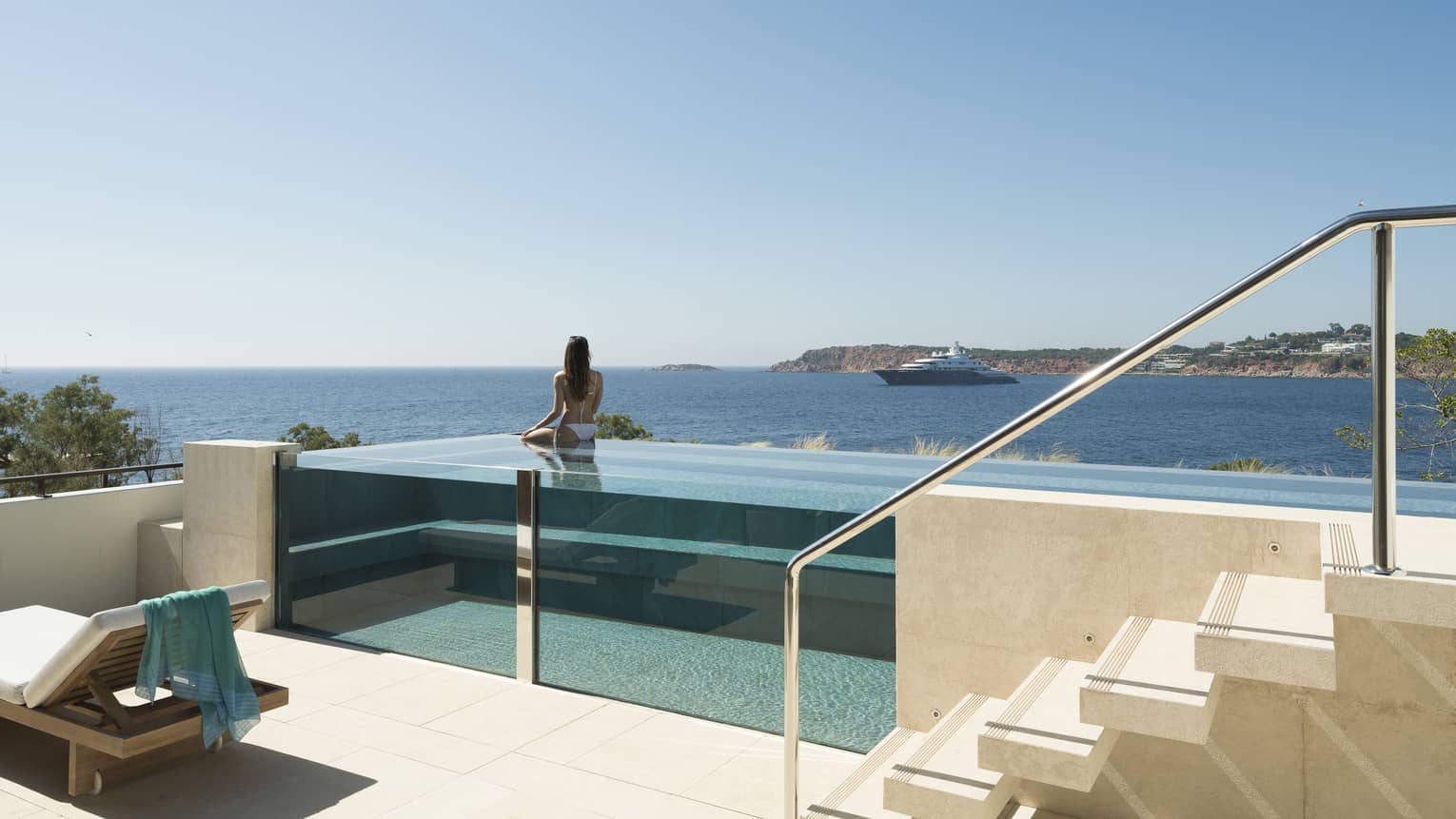 Terrace with staircase and glass infinity pool, a woman wearing white bikini in pool looks out to sea