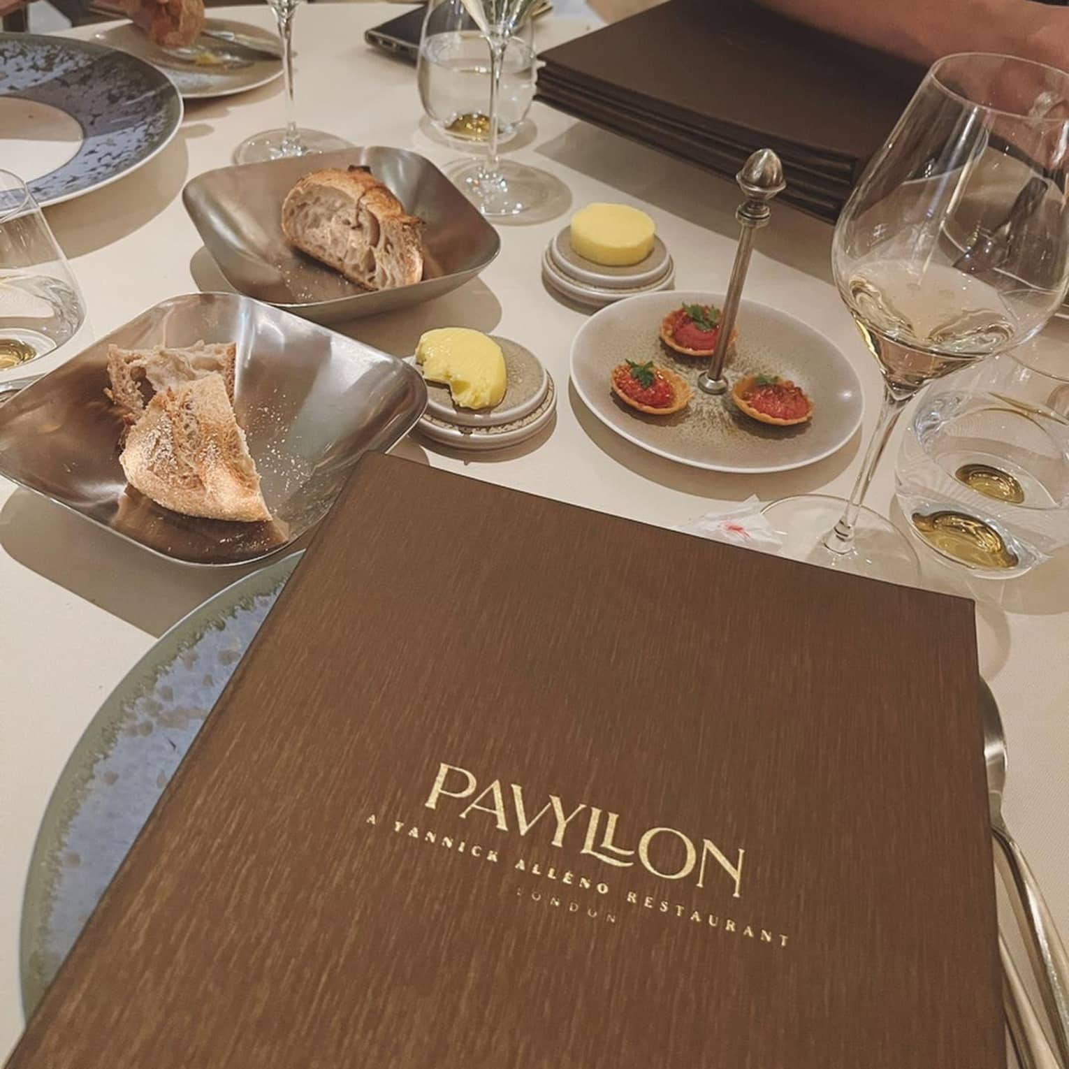 ,A brown Pavyllon menu sits on a table filled with bread, wine and hors d'oeuvres